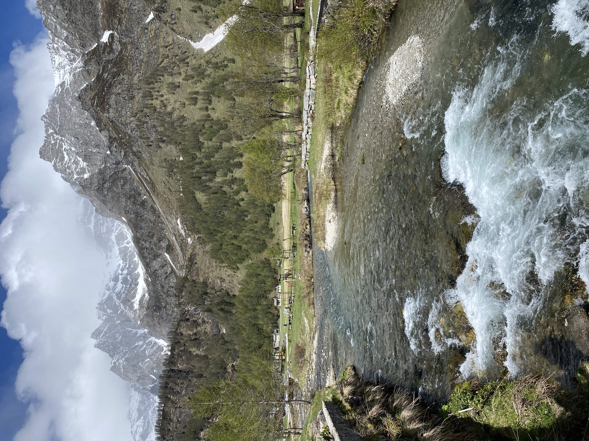 The image captures a scenic view in Baceno, Italy. A rushing river dominates the right side of the frame, its water cascading over rocks, creating white foamy rapids. To the left of the river is a grassy area with trees lining the riverbank. Further left, a dense forest climbs up the side of a massive mountain, its peak shrouded in clouds and dusted with snow. A clear blue sky is partially visible above the mountain. In the grassy area, picnic tables and other structures can be seen, suggesting a recreational space. The overall scene suggests a mountain landscape with a river running through a park area near a town, with snowy mountain peaks and forest along the park.