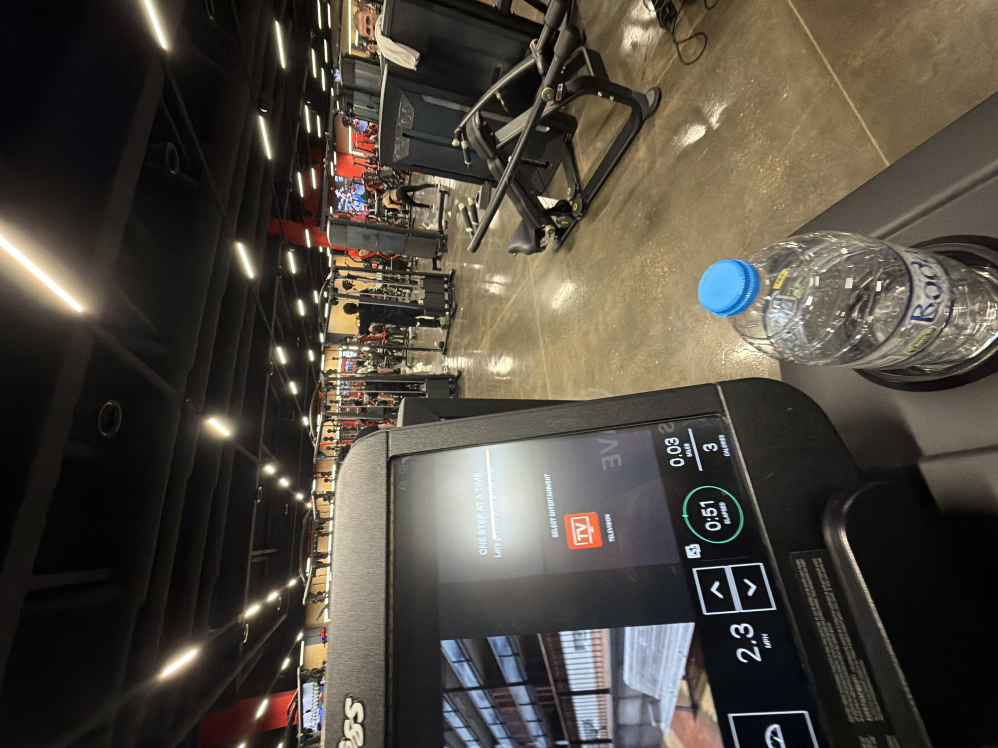 The image depicts a gym interior with a treadmill in the foreground. The treadmill's console displays "2.3 MPH," "0:51 ELAPSED," "0.03 MILES," and "3 CALORIES." A water bottle rests in a holder on the console. In the background, rows of exercise machines are visible, extending into the depth of the gym. Overhead lights illuminate the scene. The location cannot be identified as Plovdiv, Bulgaria based on the given image.