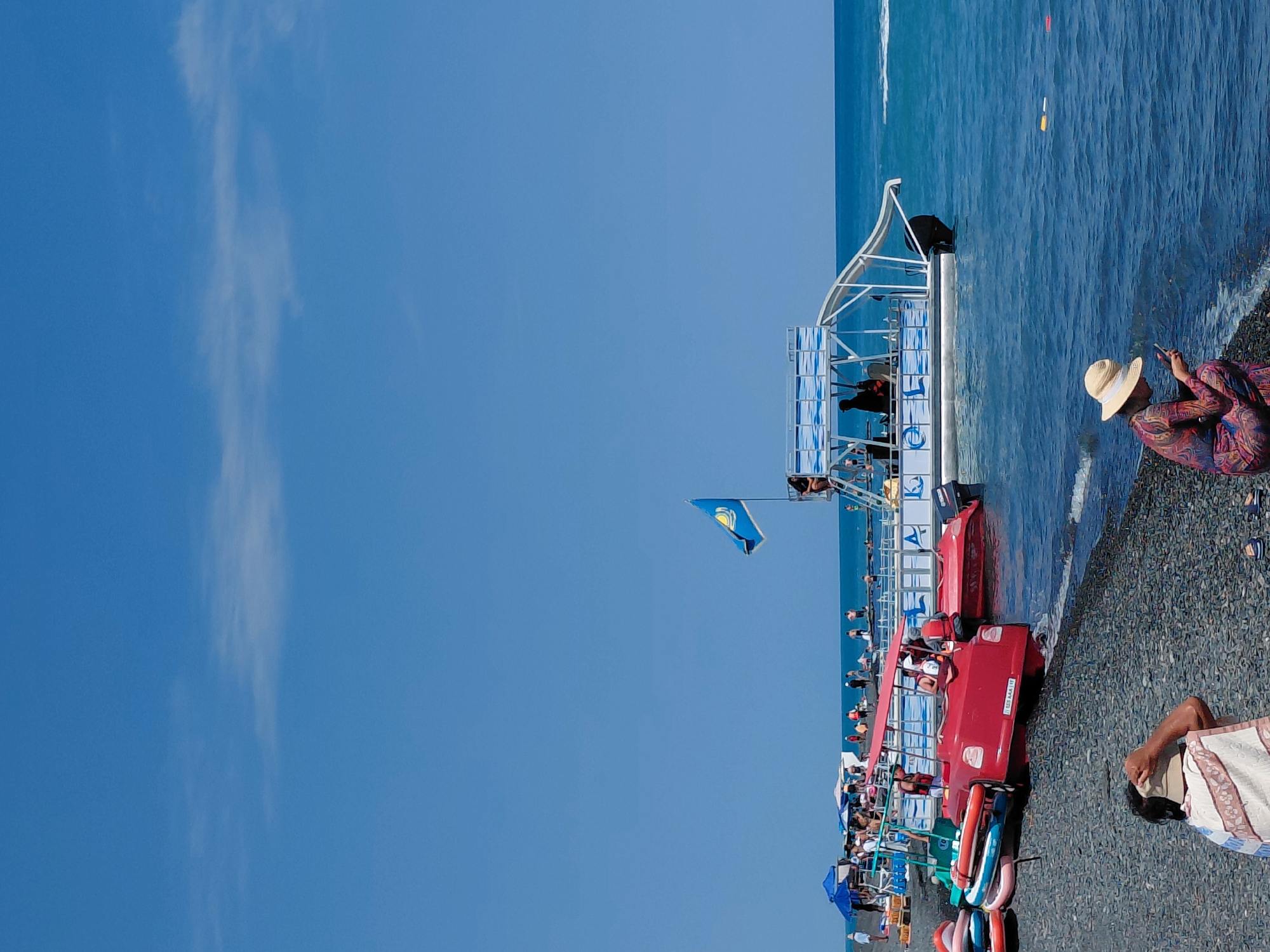 The image shows a sunny day at a pebble beach in Ақши, Kazakhstan. In the foreground, a person wearing a straw hat and a patterned dress is sitting on the beach, looking at their phone. Behind them, several colorful inflatable tubes are stacked on the shore next to a red boat. Further out in the water, a white pontoon boat with a slide is docked, with several people visible on its deck. A blue flag with a yellow emblem flutters from a pole on the boat. In the background, more people are scattered on the beach and in the water, with beach umbrellas providing shade. The ocean is a vibrant blue, and the sky is clear.