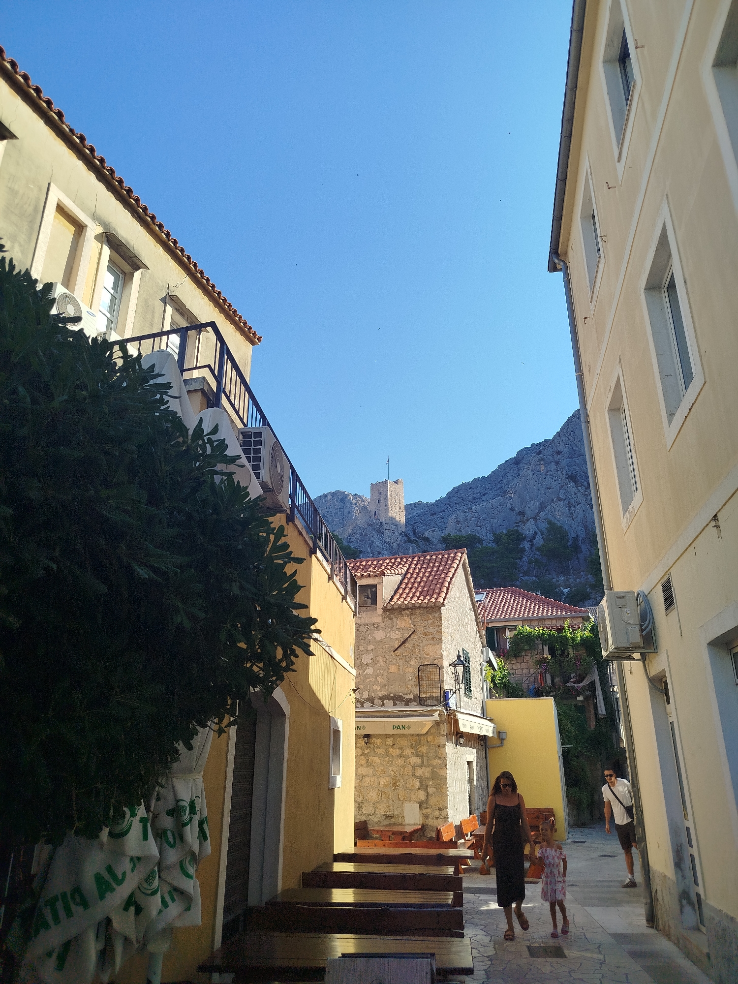 The image depicts a narrow street scene in a city with old stone buildings and a clear blue sky. On the left, a yellow building with a black railing balcony is visible, partially obscured by dark green foliage and white umbrellas with green lettering. A portion of an air conditioning unit is mounted on the building. In the center of the image, a woman in a dark dress and a young girl in a patterned dress are walking away from the camera. Behind them, tables and benches of an outdoor seating area are arranged. Further back, stone buildings with red tiled roofs are visible, and a large rocky mountain with a medieval tower on top dominates the background. On the right, a light-colored building with windows and an air conditioning unit is seen. Another person, a man wearing a white t-shirt and shorts, is walking towards the foreground on the right side of the street. The scene appears to be bathed in sunlight, casting shadows on the ground.