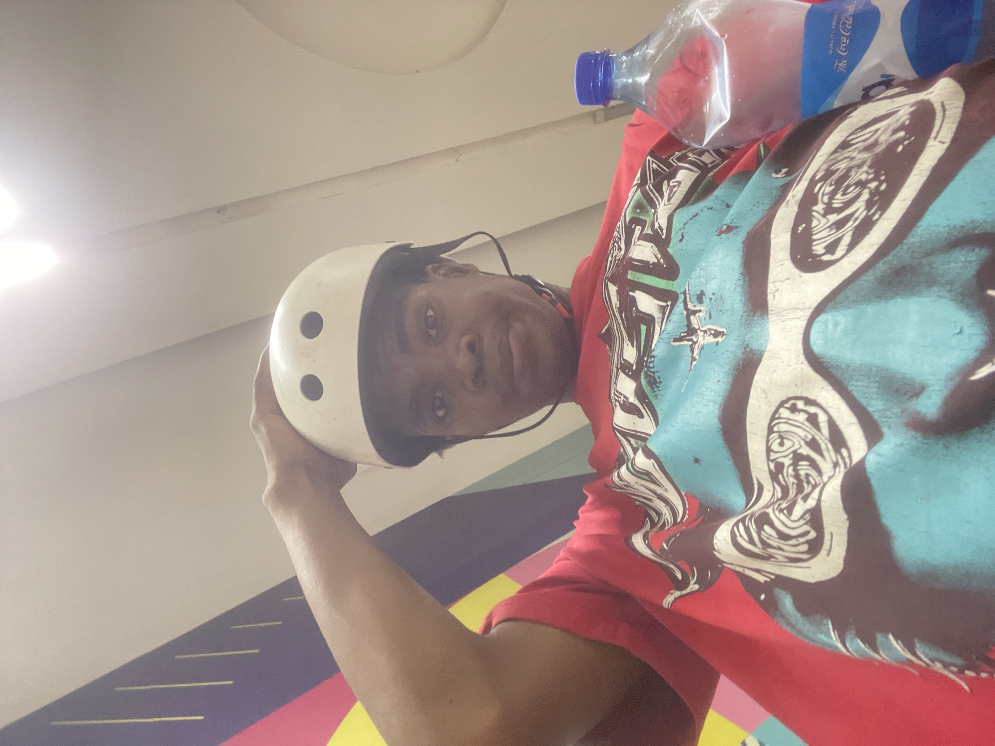 A low-angle, medium shot shows a person wearing a white helmet and a red t-shirt with a graphic design. The person's right arm is raised, holding the helmet. A clear plastic bottle of Coca-Cola is visible to the right of the person. The background features a colorful, abstract mural. The scene appears to be indoors, possibly in Moba, Nigeria. The lighting is bright, suggesting overhead illumination.