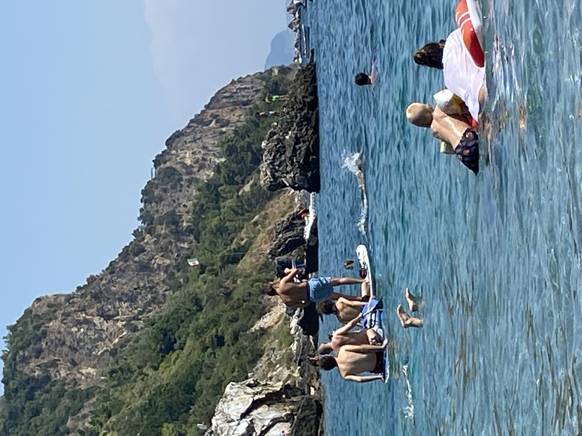 This image depicts a sunny day at a coastal location with a rocky cliff face in the background. The water is clear and blue, with several people engaged in various water activities. In the foreground, two children are sitting on inflatable toys in the water, facing away from the camera. To the left, several people are on a paddleboard, with one person standing. Further back, a person is swimming in the water, and another is visible on a rocky outcrop on the cliff. There are also several boats anchored in the distance. The cliff face is covered in lush green vegetation, and a small flag is visible on its side. The overall scene suggests a recreational day at the beach or bay, likely in a Mediterranean or similar climate. The prompt specifies Prijevor, Montenegro, as the location.