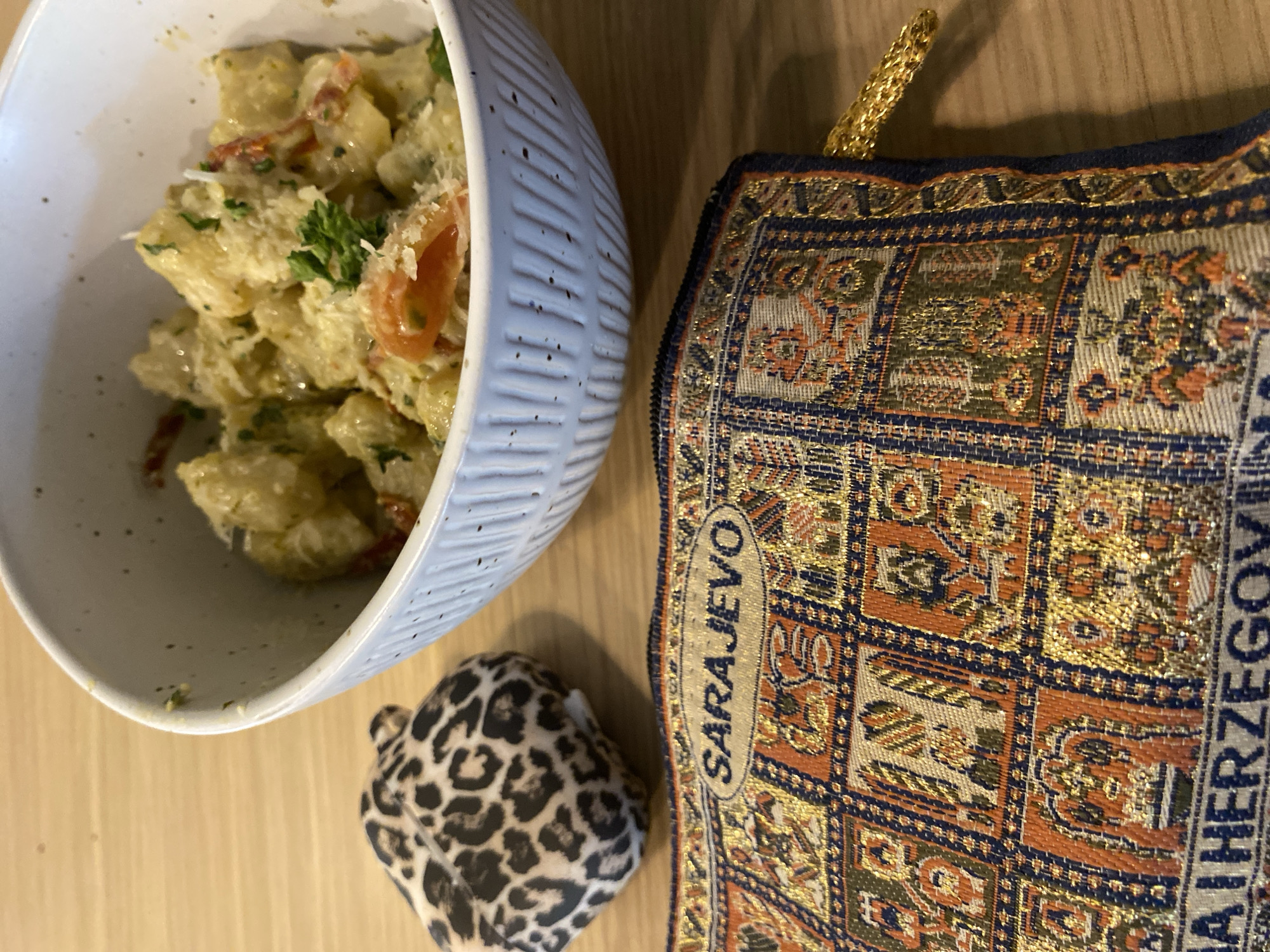 The image shows a bowl of food next to a leopard print pouch and a fabric souvenir bag labeled "Sarajevo." The bowl, containing what appears to be pasta or potato salad with tomatoes and herbs, is light blue with vertical ridges. The fabric bag has a gold handle and intricate patterns in gold, orange, and blue. All the items are arranged on a wooden table. There is no discernible indication that the photo was taken in Novi Sad, Serbia.