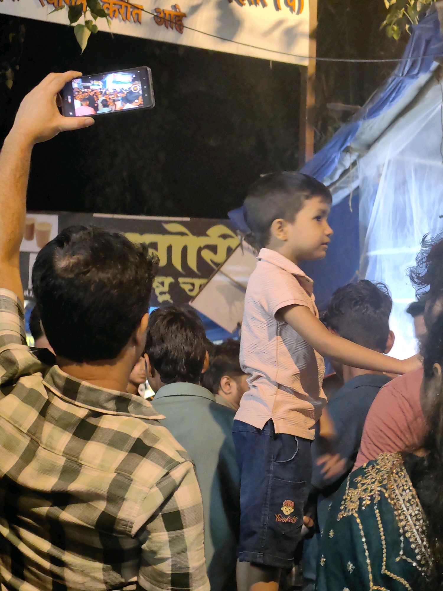 In Mumbai, India, a person is holding a smartphone horizontally, capturing a scene with a crowd. In the foreground, a person in a plaid shirt is visible from the back, with their left arm raised holding the phone. A young boy in a pink collared shirt and dark shorts stands in the middle ground, looking to the right. Behind him, other individuals are seen, including a person in a green shirt and another in a dark blue shirt. The background features a banner with text in Marathi and what appears to be a tent-like structure with blue and white fabric. The lighting suggests it is evening or nighttime, with artificial lights illuminating the scene. The overall atmosphere is that of an outdoor gathering or event.