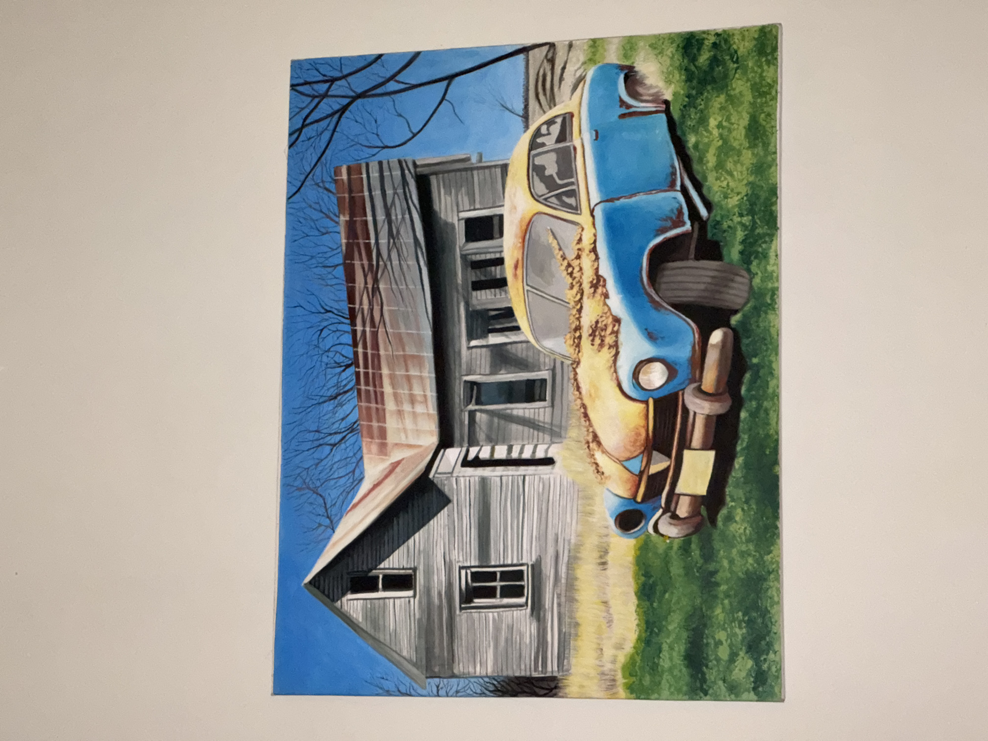 The image is a painting of a dilapidated barn with a rusted, vintage car parked in front of it. The barn is grey with a rusted roof and several windows. Bare branches of trees are visible against a blue sky in the background. The car is predominantly blue and yellow with extensive rust and damage. It is situated on a patch of green grass. The painting appears to be hanging on a plain white wall. There is no evidence suggesting the location is in Kuta Selatan, Indonesia.