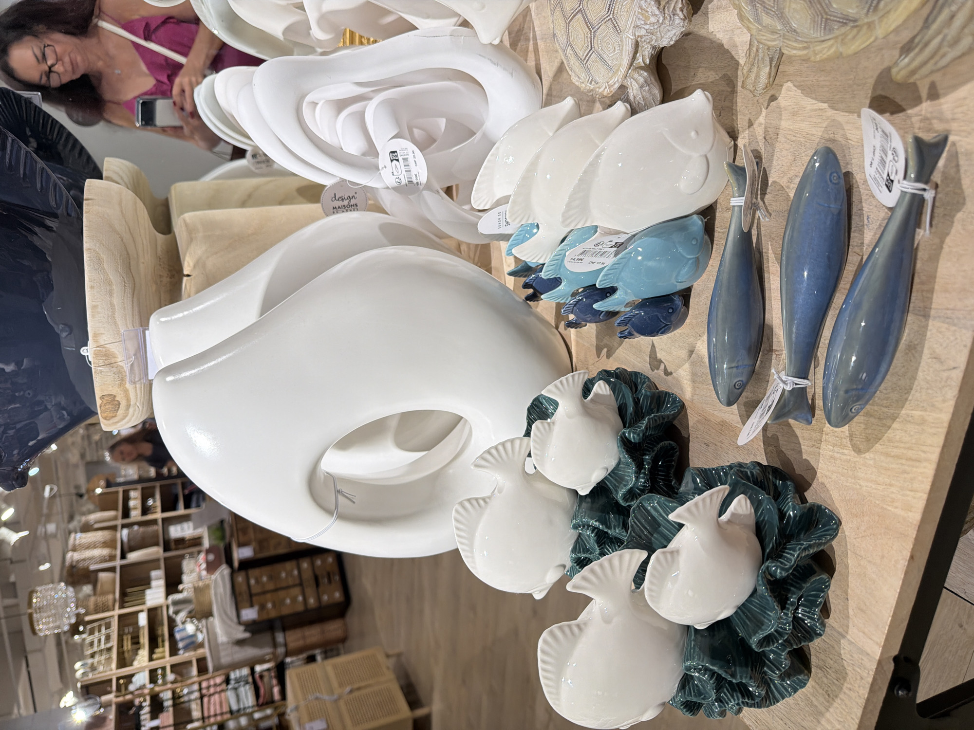 An indoor display of decorative home goods is shown. In the foreground, a collection of white ceramic fish sculptures with green bases are arranged on a wooden surface. To the right of the fish, several elongated, blue ceramic fish sculptures with fins are positioned, some with price tags attached. Behind these, a large, white, abstract ceramic sculpture is visible, along with other white sculptural elements. In the background, shelves stocked with various items, including baskets and what appear to be wooden drawers, are visible. A woman wearing a pink top and glasses is standing in the background, holding a smartphone. The overall environment suggests a retail setting.