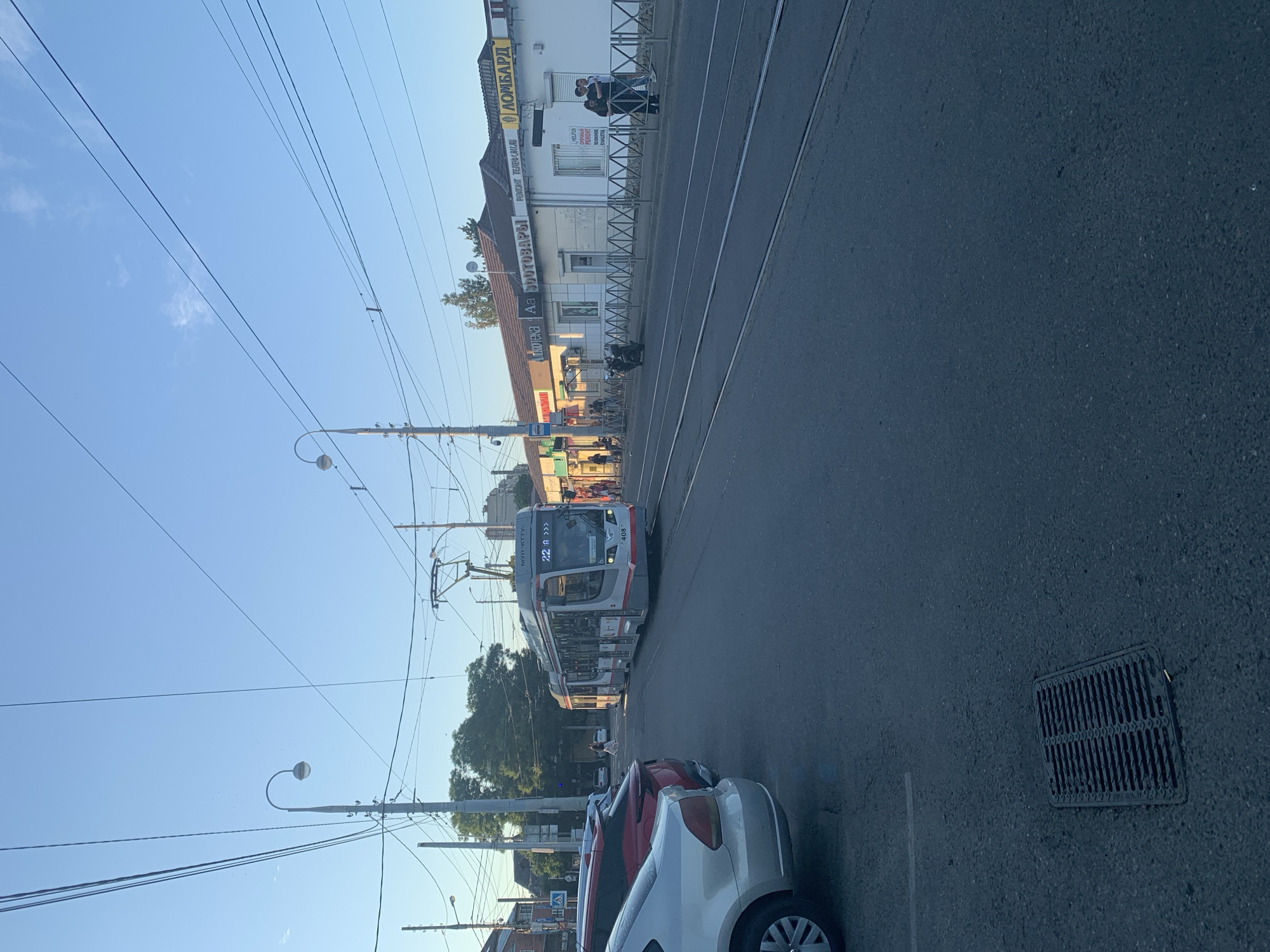 The image depicts a street scene in a city. A modern tram, numbered 408 and displaying route "22 A >>>", is traveling on tram tracks. In the foreground, the left side of the image shows the side of a silver car, and a red car is parked behind it. A drainage grate is visible on the asphalt in the lower left corner. On the right side of the street, a row of storefronts is visible, with signs in Russian indicating businesses such as "Ломбард" (Pawnshop), "РЕМОНТ ТЕЛЕФОНОВ" (Phone Repair), and "ОБУВЬ" (Shoes). Several people are seen walking and standing near the storefronts and on the sidewalk. Overhead, a network of power lines and streetlights crisscrosses the sky, which is clear and blue. Trees are visible in the background behind the buildings. The overall scene suggests a typical urban environment during daylight.
