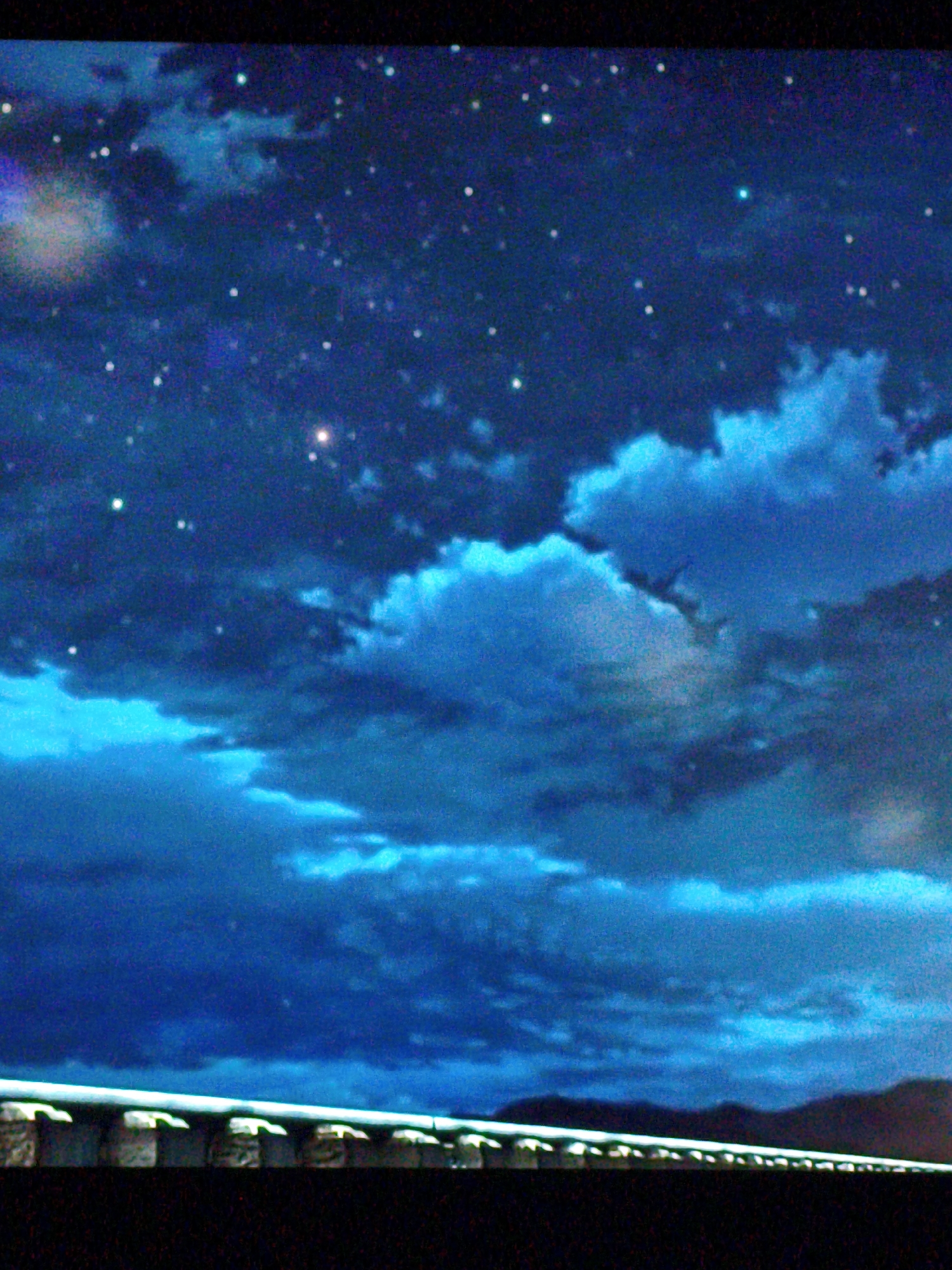 The image captures a nighttime view of a bridge in Vladivostok, Russia, set against a backdrop of a starry sky and illuminated clouds. The bridge, likely an iconic landmark of the city such as the Golden Horn Bay Bridge, stretches horizontally across the lower portion of the frame. It is brightly lit, with rows of lights casting a glow upwards onto the underside of the bridge's structure. The bridge's concrete pylons are visible, each supporting the illuminated deck. In the distance, faint silhouettes of hills or mountains can be seen against the horizon. The sky is a deep blue, speckled with numerous stars and adorned with large, luminous clouds that appear to be lit from above, possibly by the moon or city lights. The overall scene conveys a serene and somewhat surreal atmosphere, characteristic of a clear night in a coastal city.