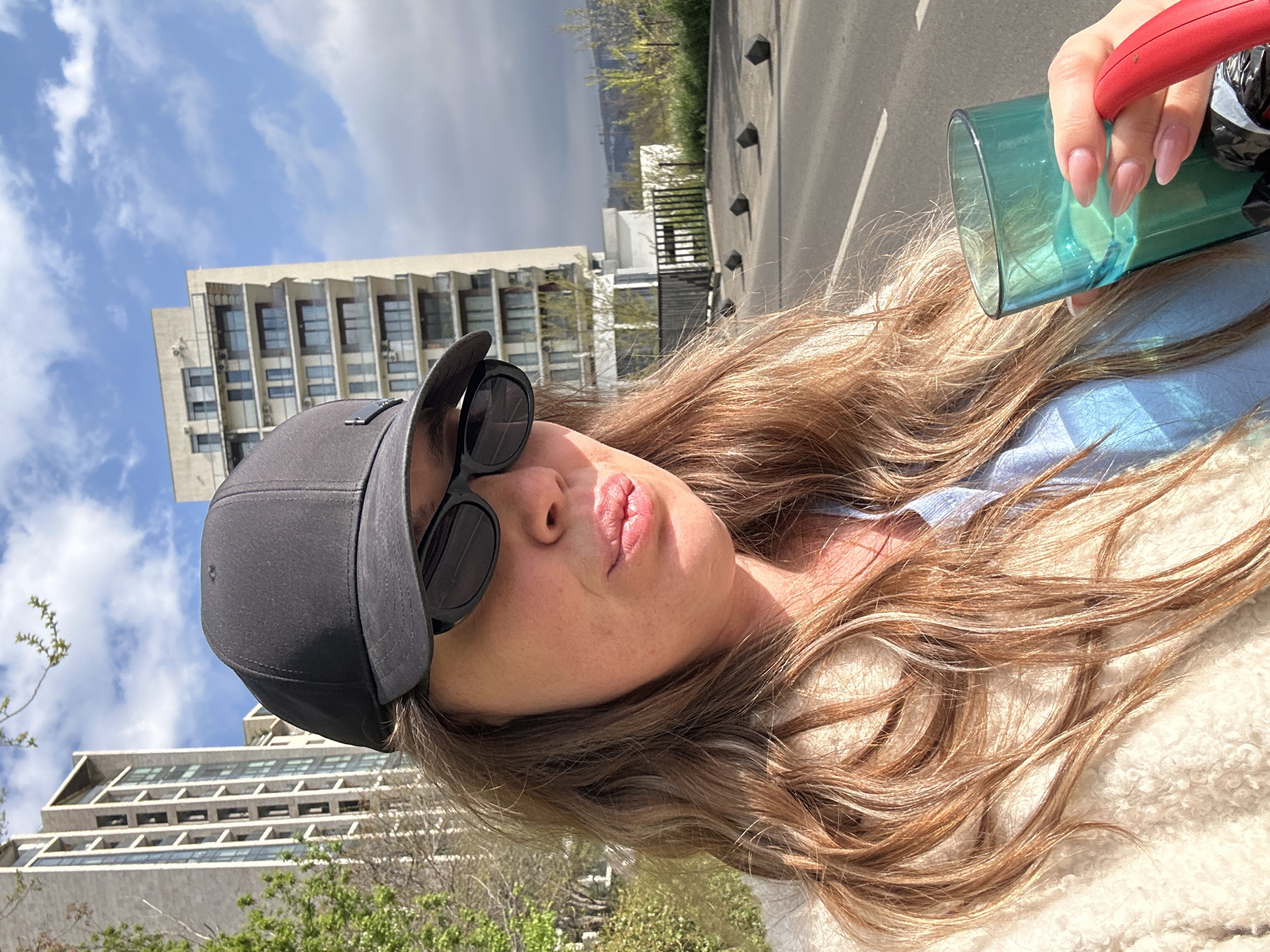 A woman with long blonde hair, wearing a black baseball cap and sunglasses, poses for a selfie. She is holding a green glass with a red handle, against a backdrop of buildings and a cloudy sky. The location is Tbilisi, Georgia, as identified by the distinctive architecture of the buildings. The street is visible on the right with bollards.