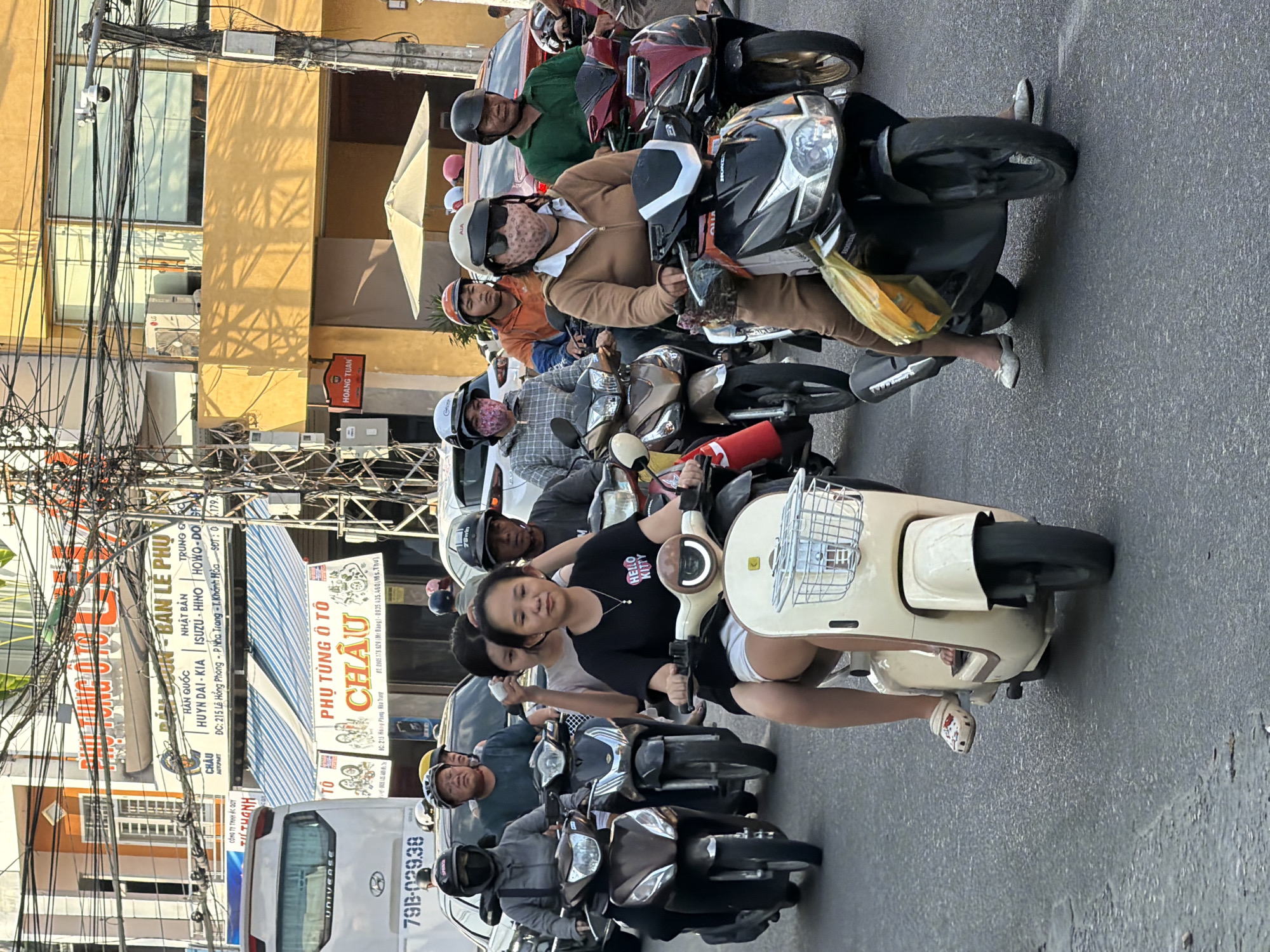 The image captures a street scene in Nha Trang, Vietnam, likely during daytime. A procession of motorbikes navigates a busy street, suggesting a typical urban traffic scenario. In the foreground, several individuals are riding motorbikes. One woman in the front, wearing a black t-shirt with a "Hello Kitty" graphic and white shorts, is riding a light-colored scooter with a basket at the front. She is looking forward with a neutral expression. To her right, another woman in a tan tracksuit and a patterned face mask rides a black motorbike. To the left of the primary subject, a woman in a grey jacket and a patterned face mask is on a dark brown scooter, and behind her, a person in a green shirt is also on a motorbike. The background reveals buildings with shops, including signs in Vietnamese indicating automotive parts ("PHỤ TÙNG Ô TÔ CHÂU"). Electrical wires are prominently strung overhead, a common sight in Vietnamese cities. A white bus with "UNIVERSE" printed on its side and a license plate starting with "79B-" is visible on the left. The street surface is asphalt. The overall atmosphere appears to be one of active urban movement.