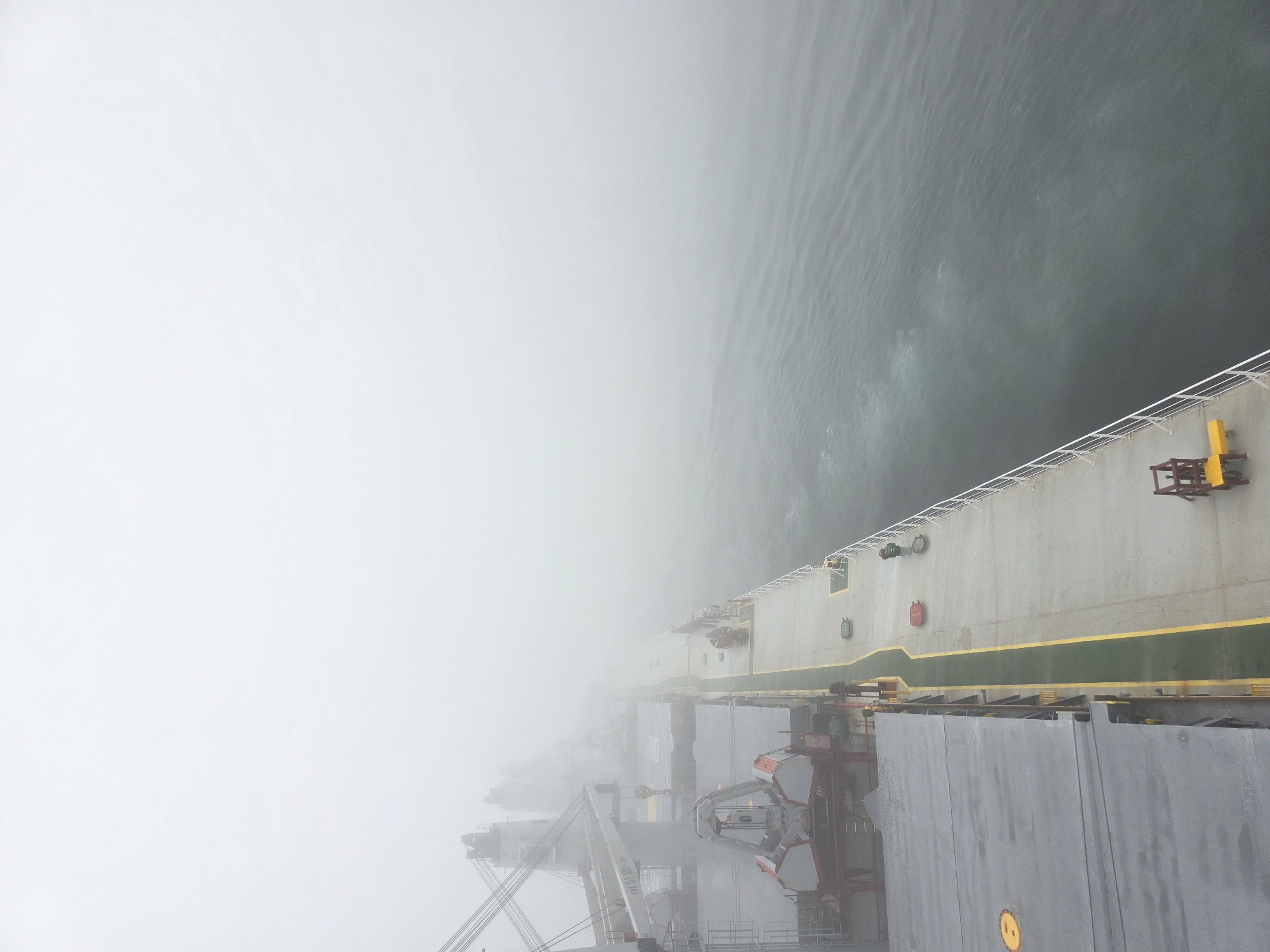 The image depicts the deck of a large cargo ship on a foggy day. The ship's deck is predominantly gray with yellow and green markings. Several large cranes are visible in the background, partially obscured by the fog. On the deck, there are various pieces of equipment, including what appear to be lifeboats and a yellow industrial cart. The water beside the ship is choppy and dark, with visible ripples. The overall atmosphere is hazy and diffused due to the dense fog. There are no people clearly visible on the deck. The location is given as Unknown, Unknown.