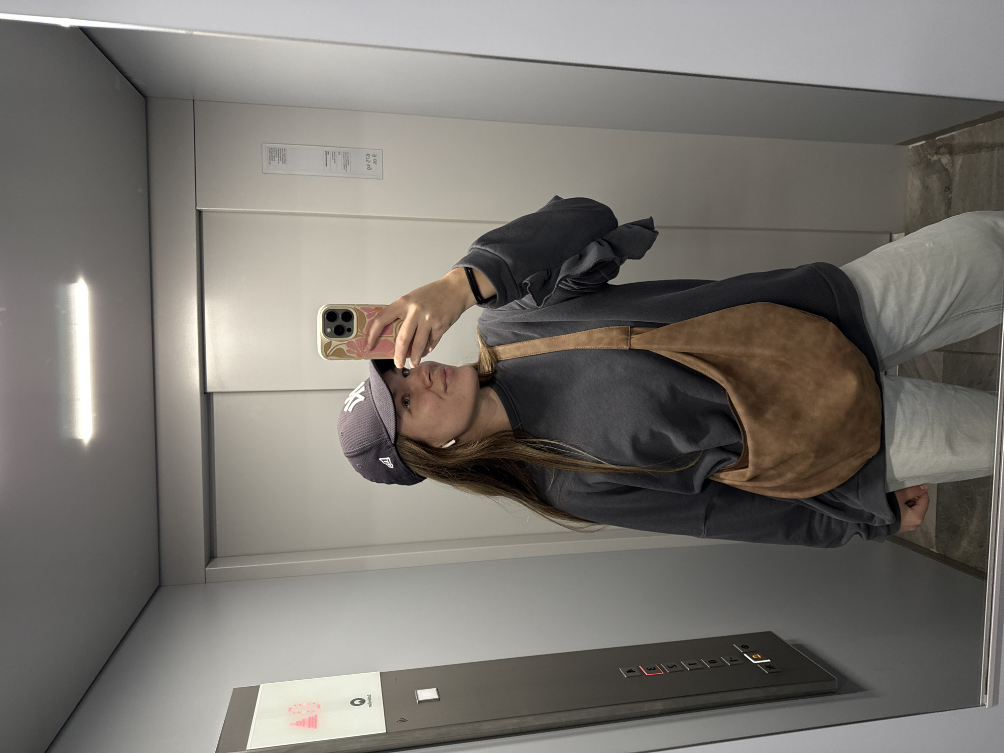 A young woman is taking a selfie in an elevator. She is wearing a dark grey sweatshirt, light grey jeans, a grey baseball cap with the NY logo, and a brown shoulder bag. She holds her phone in her right hand. The elevator has a grey control panel with illuminated floor numbers. The walls of the elevator are light grey. The elevator is located in Vozdovac Urban Municipality, Serbia.