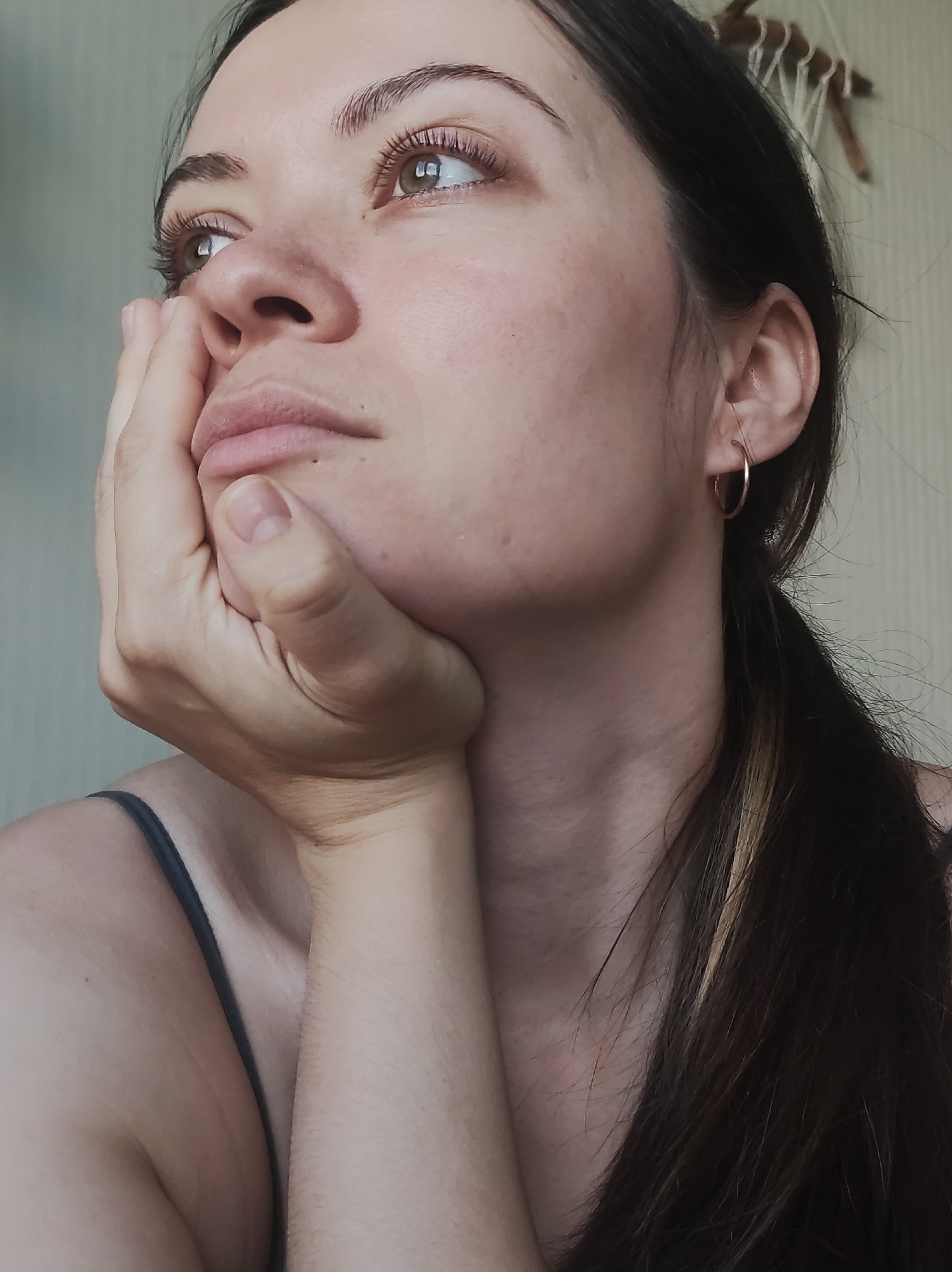 A close-up, head-and-shoulders portrait captures a woman looking upwards and slightly to her left with a thoughtful or pensive expression, likely taken indoors in Chaikovsky, Russia. Her right hand supports her chin and lower jaw, with her fingers gently curled.
The woman has fair skin, with visible freckles on her cheek and forehead. Her olive-green eyes are open and directed upwards, framed by long, dark eyelashes and dark, well-defined eyebrows. Her lips are slightly parted. Her dark hair is pulled back, possibly into a low ponytail, revealing a streak of lighter, possibly dyed, hair near her neck. A simple gold hoop earring is worn in her right ear. She is wearing a dark, thin-strapped top or camisole, visible on her left shoulder and neckline.
The background is a plain, light-colored wall. A decorative, woven item made of natural fibers or wood is partially visible at the top right corner, hanging on the wall. The scene is illuminated by soft, natural light, suggesting it is daytime. There is no visible text in the image.
