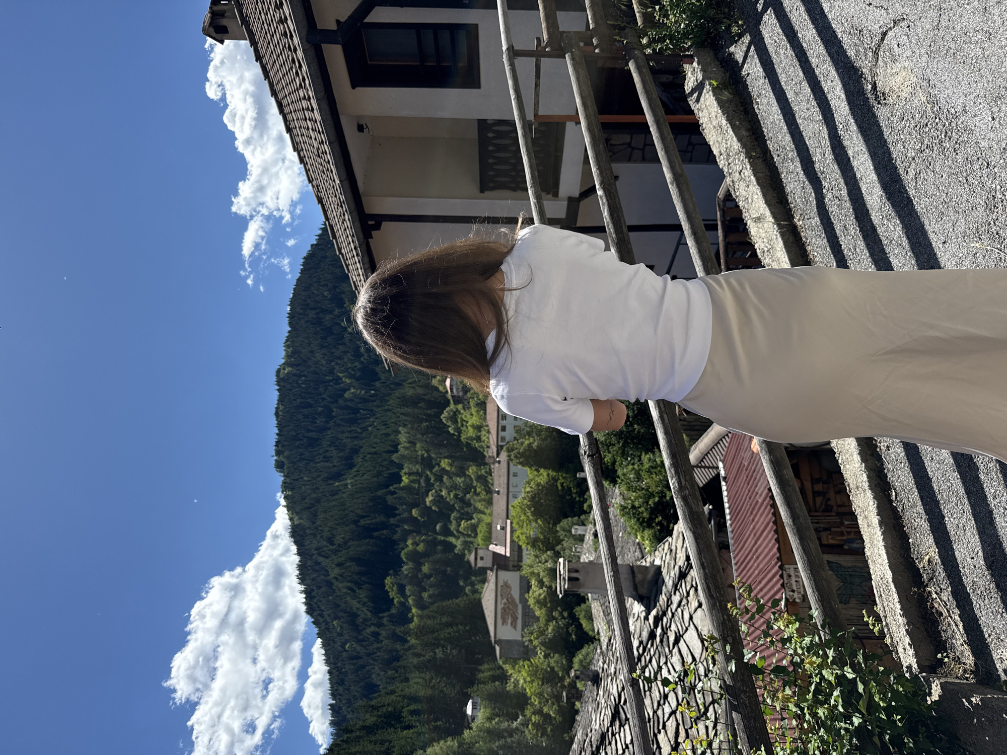A person with long, brown hair stands with their back to the camera, looking out over a village nestled in a valley. They are wearing a white t-shirt and a long, beige skirt. In the foreground, there is a wooden railing and a stone wall. Behind the person, the village of Shiroka Laka, Bulgaria, is visible, with traditional stone houses and red-tiled roofs. The houses are surrounded by lush green trees, and a dense forest covers the mountainside in the background. The sky is a clear blue with a few scattered white clouds. The scene suggests a peaceful, rural setting on a sunny day.