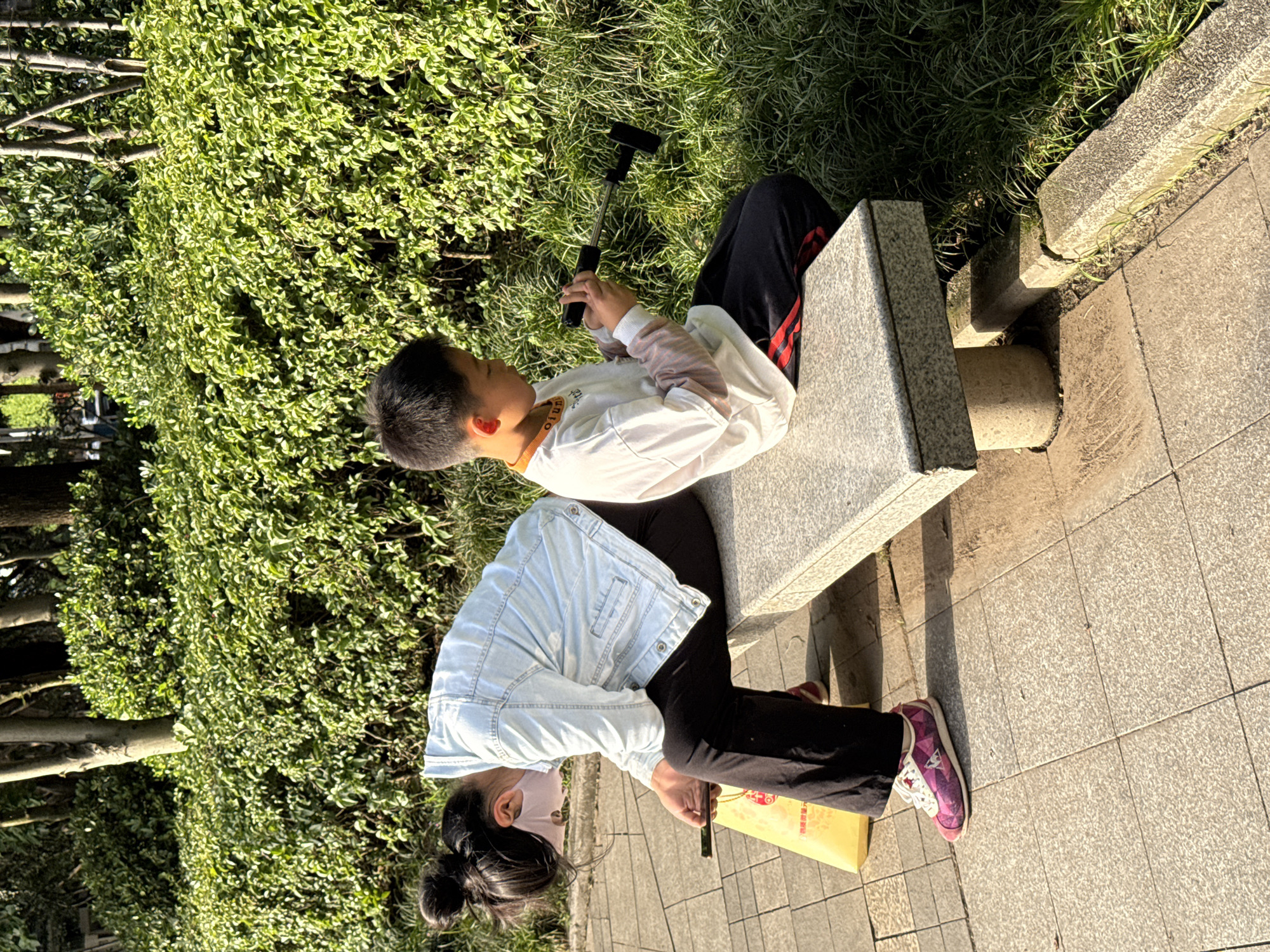 An outdoor scene in Jiangjin, China, depicts two young individuals sitting on a grey stone bench in a park or public green space. The image is taken on a sunny day, likely in the late morning or late afternoon, as indicated by the direct sunlight and distinct shadows.
On the right side of the bench sits a boy with short dark hair. He is wearing a white long-sleeved shirt with "olun" visible on the front, black athletic pants with red stripes down the side, and a gold-colored lanyard around his neck. He holds a black monopod or selfie stick in both hands, intently looking at a smartphone attached to its end, suggesting he is filming, vlogging, or taking photos.
On the left side of the bench, closer to the viewer, sits a girl or young woman. Her dark hair is tied back in a bun, and she is wearing a light pink face mask. She is dressed in a light blue denim jacket over a dark top, black pants, and distinctive pink and white geometric-patterned sneakers with white laces. She holds a smartphone in her right hand, seemingly engrossed in it. A yellow paper bag with some red Chinese characters (not clearly legible) rests on the tiled ground beside her.
The bench is situated on a grey tiled pathway, bordered by lush green bushes with small, variegated leaves directly behind it. Beyond the bushes, taller trees are visible, reinforcing the park-like setting. The ground surrounding the bench also features patches of green grass. Both individuals appear calm and focused on their respective devices, enjoying the outdoor environment.