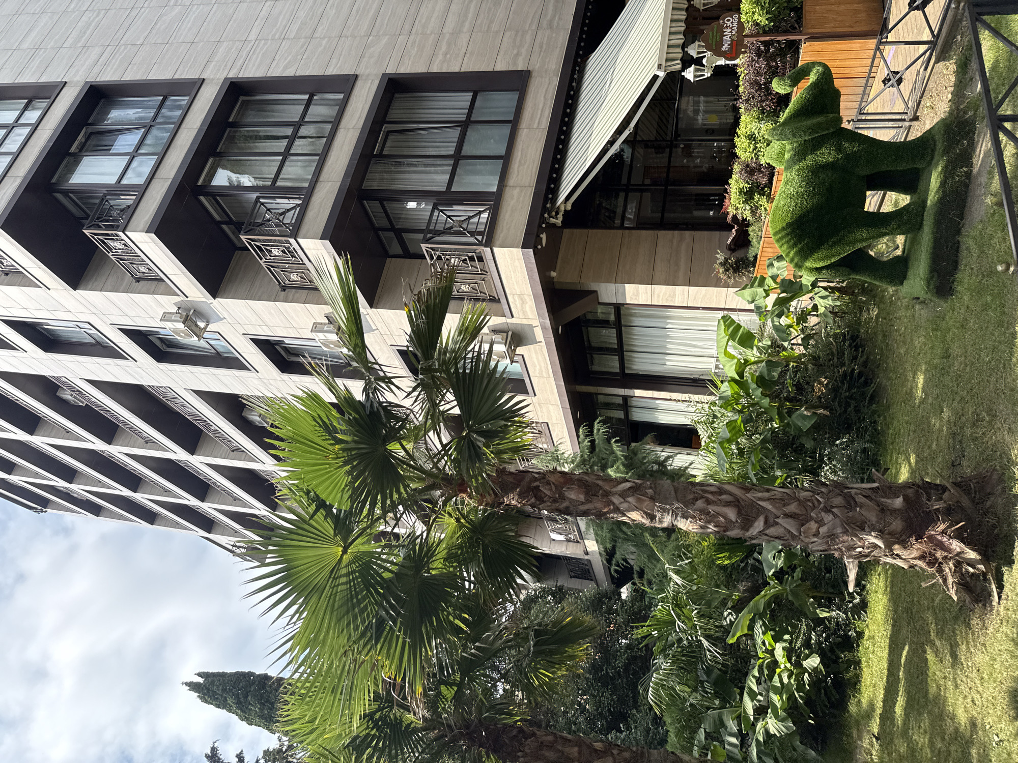 In the city of Sochi, Russia, a modern, multi-story hotel building with beige tiled facade and dark balconies stands tall. In the foreground, lush green palm trees and dense tropical foliage create a vibrant garden setting. To the right, a whimsical, topiary elephant, sculpted from green foliage, stands on a grassy mound, adding a playful touch to the scene. The environment suggests a sunny day with a partly cloudy sky, indicative of a warm climate.