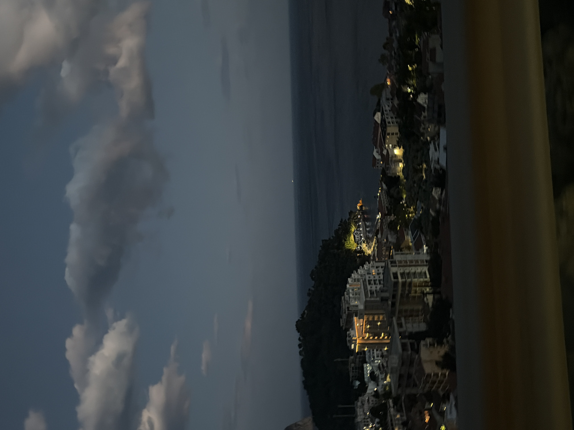 An aerial view from a high vantage point overlooking the city of Nerin, Montenegro at night. The sky is filled with clouds, with the sea visible in the center of the frame. The city is lit up with artificial light. A large cylindrical object is visible in the foreground.