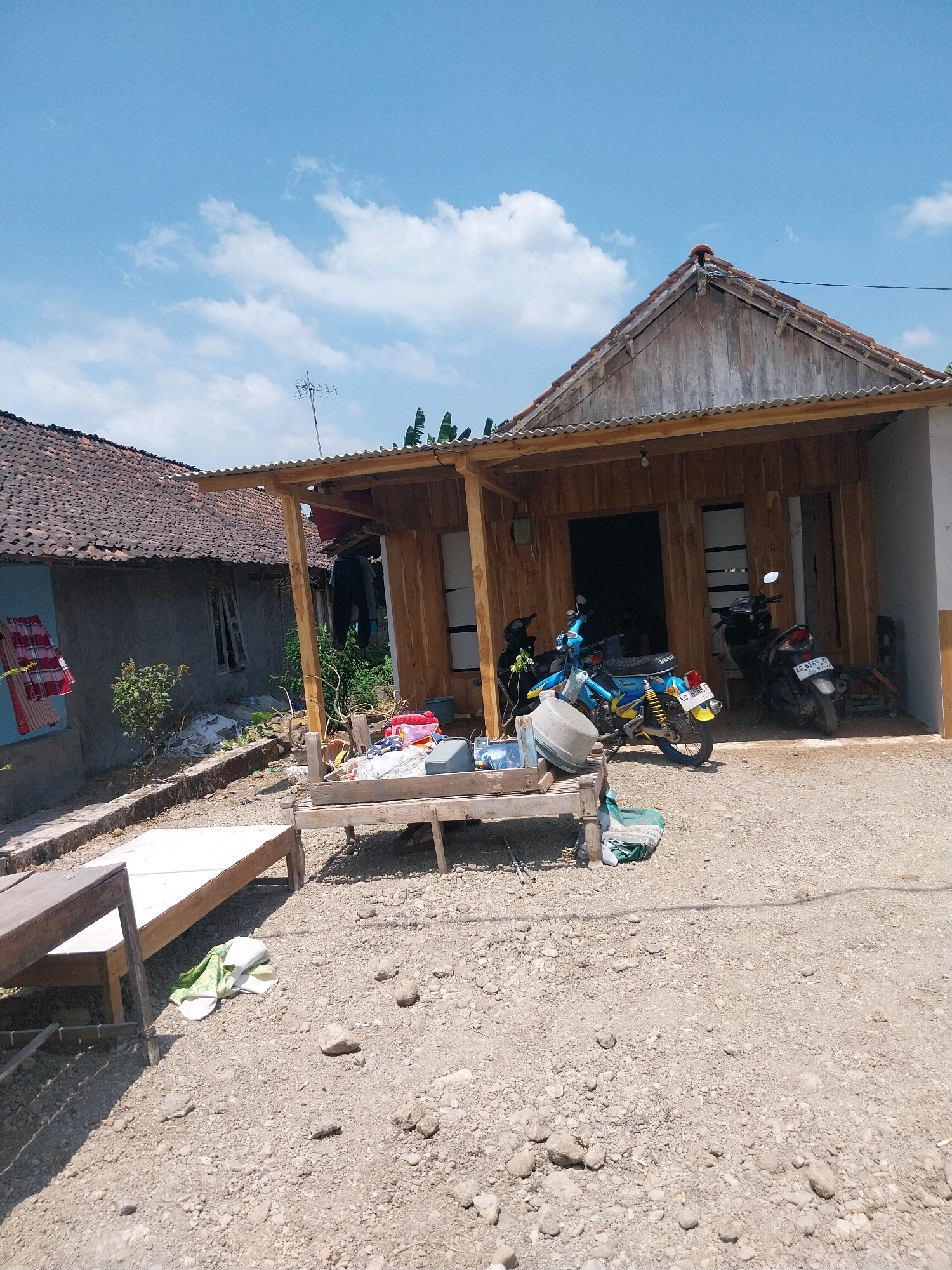 The image depicts an outdoor scene in a rural area, possibly the city of Campur, Indonesia. In the foreground, there is a gravel-covered yard with two wooden benches. A green cloth is draped over one of the benches. Behind the benches is a partially constructed wooden building with a corrugated metal roof. Two motorcycles are parked in front of the building: a blue and yellow motorcycle on the left and a black motorcycle on the right.
To the left of the wooden building, there is a darker, older building with a tiled roof. Clothes are hanging on a line on the side of this older building. In the background, there are trees and a bright blue sky with some white clouds. The overall impression is of a humble, rural dwelling under construction.