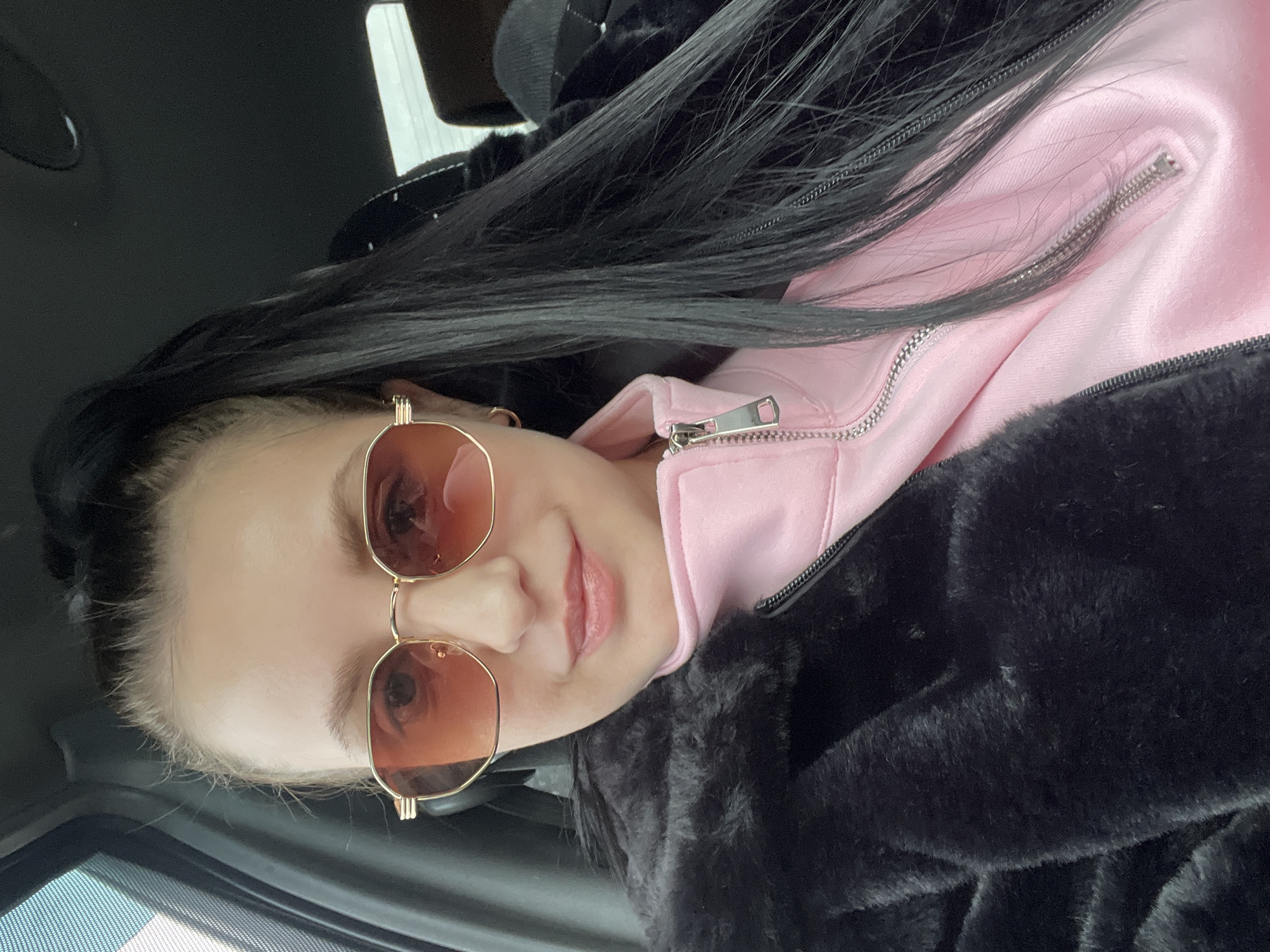 An individual, identified as female, is depicted in a close-up, chest-up selfie taken inside a vehicle in Chaikovsky, Russia. She has fair skin, dark hair styled in a high ponytail, and is wearing gold-framed sunglasses with orange-tinted lenses. Her mouth is slightly open with a light pink lip color visible. She wears small gold hoop earrings. Her attire consists of a light pink, zippered top with a silver-colored zipper pull, underneath a black faux fur jacket. The vehicle's dark gray interior is visible in the background, including a window frame on the left and a portion of the headliner above. The subject is looking directly at the camera.