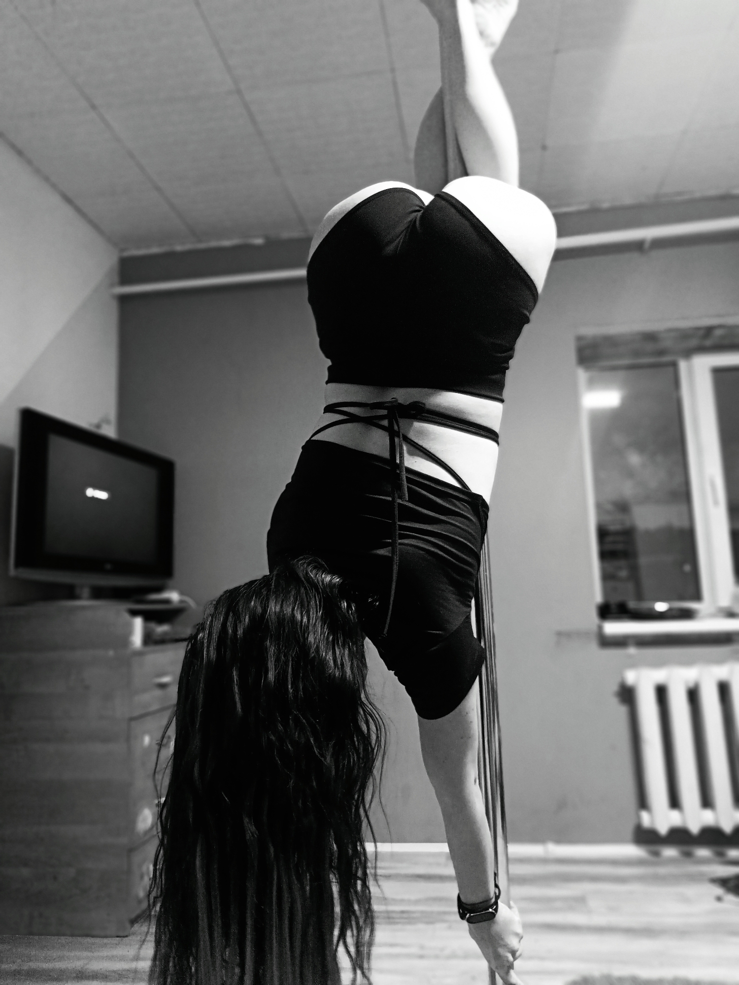 A black and white image captures a woman upside down on a pole, presumably practicing pole dancing, within an indoor setting.
The woman is positioned in the center of the frame, her legs wrapped around the pole above her head. She wears black shorts and a top, and her long dark hair cascades downwards towards the floor. A wraparound detail accents her midsection. Her hand grips the pole near the bottom.
Behind her is a room with a television set on a dresser to the left. A window is visible on the right side of the background. The room seems sparsely furnished.
The location is Unknown, Unknown.