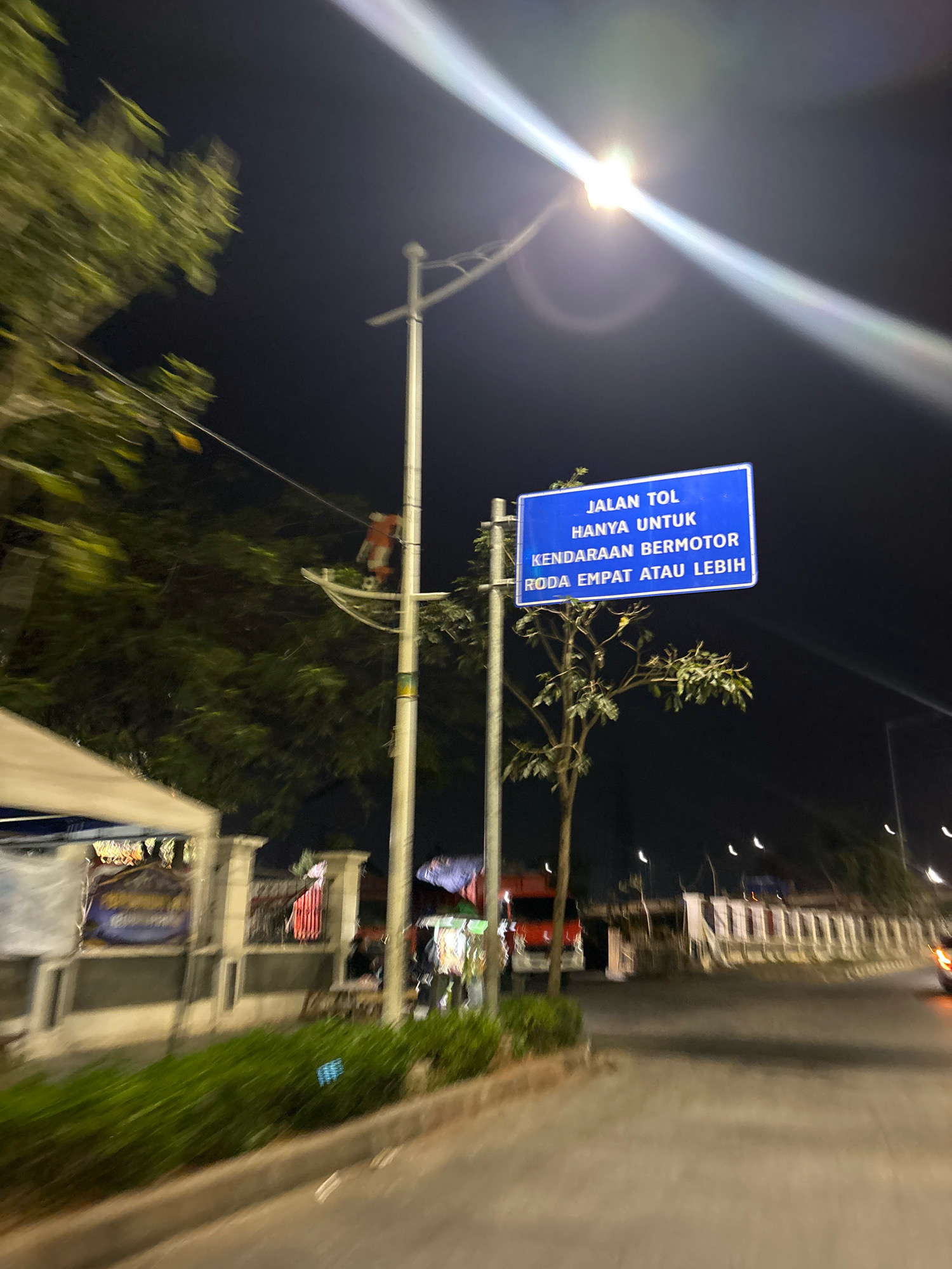 The image captures a street scene at night in North Jakarta, Indonesia, likely near a highway entrance. The focal point is a blue rectangular sign with white text indicating "JALAN TOL HANYA UNTUK KENDARAAN BERMOTOR RODA EMPAT ATAU LEBIH," which translates to "Toll Road Only for Four-Wheeled Motorized Vehicles or More." This sign is mounted on a pole illuminated by a bright street light, causing a significant lens flare that streaks across the upper part of the image.
To the left of the sign, a tree with green foliage is visible, its leaves slightly blurred due to motion. Further to the left, a white tent structure and a fence line are present, suggesting a roadside market or event. A traffic light or street lamp pole stands tall, with what appears to be a draped orange object and a soccer ball suspended from a net attached to it.
The road surface is visible in the foreground and stretches into the background, with motion blur indicating movement within the scene. In the distance, other street lights and structures, including what looks like an overpass or bridge, are faintly discernible. The overall atmosphere is dark, illuminated by artificial lights, and slightly hazy. No people are clearly identifiable in the frame.