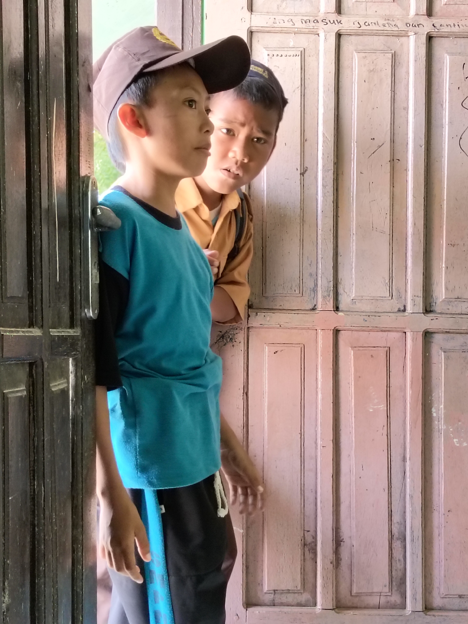 Two young boys, likely Indonesian given the location of Lori, Indonesia, are partially visible standing by a doorway. The boy in the foreground, wearing a brown baseball cap and a teal t-shirt with black sleeves, stands in profile facing left. He has short dark hair and his gaze is directed outward. To his right and slightly behind him, another boy, wearing a tan uniform shirt, looks towards the camera with a surprised or inquisitive expression. He has short dark hair. The background consists of a wooden door with a paneled design, painted a light pink color, with some visible writing in black on the upper section of the door. The boy in the foreground is positioned near the dark brown wooden frame of the doorway. The lighting suggests an outdoor or well-lit indoor environment.