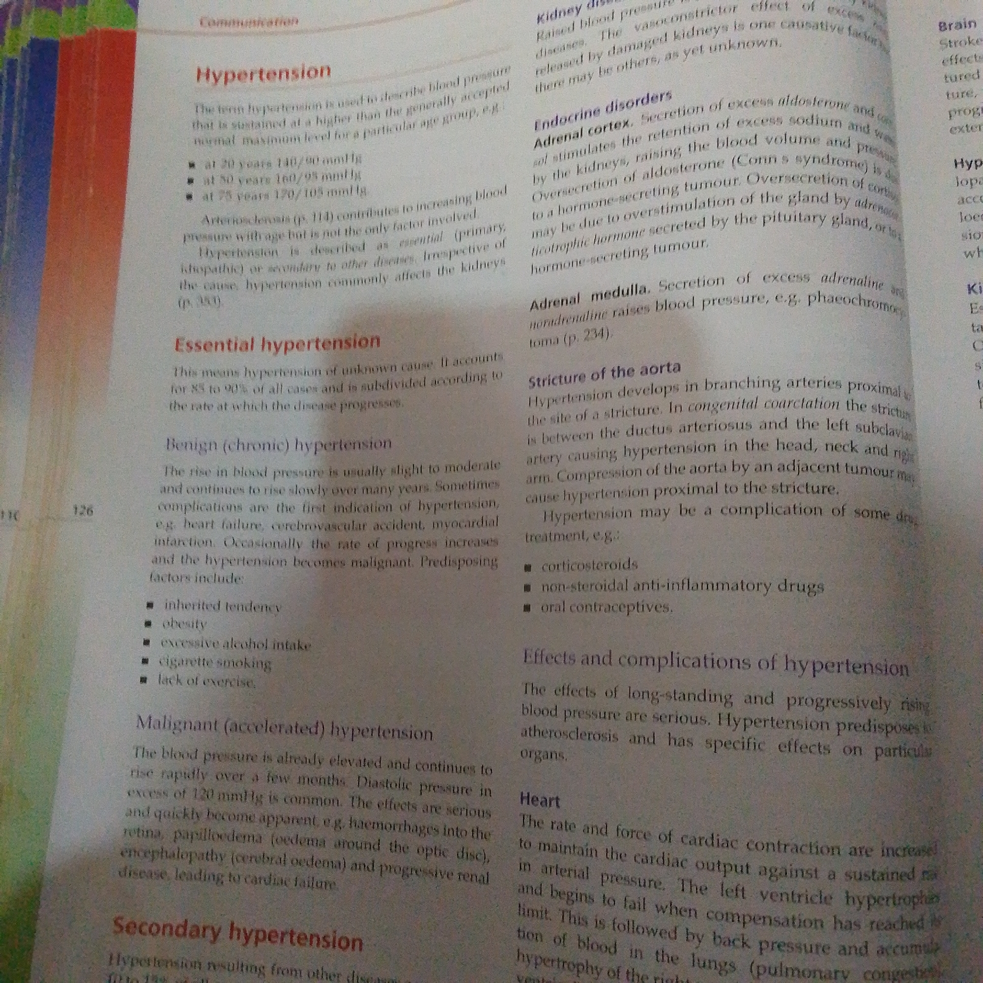The media file is a photograph of an open book with text and some colorful pages visible on the left side. The book appears to be a medical textbook, with sections on "Hypertension," "Essential hypertension," "Benign (chronic) hypertension," "Malignant (accelerated) hypertension," and "Secondary hypertension." There are also sections on "Kidney disease," "Endocrine disorders," and "Stricture of the aorta," as well as "Effects and complications of hypertension." The text discusses various aspects of high blood pressure, its causes, and its effects on the body. Visible text includes page numbers "110" and "126." The setting is likely an indoor environment, such as a study or library. No people are visible in the image. The image is well-lit, and the text is clear and readable. The colorful pages on the left suggest the book is part of a larger volume with visually distinct sections or dividers.