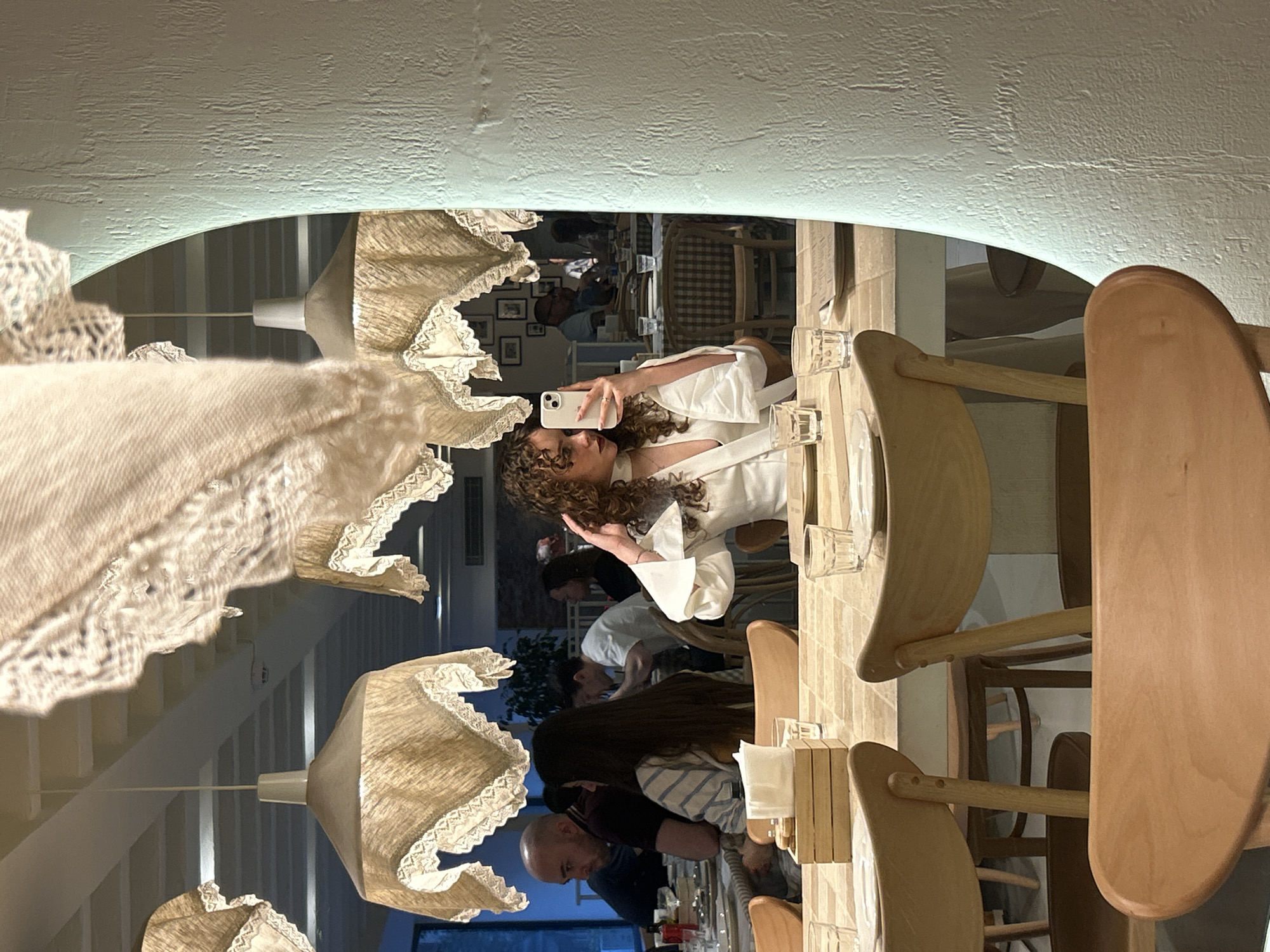 The image shows a mirror reflection of a restaurant interior, likely in Belgrade, Serbia. A woman with curly hair, dressed in a white top, is taking a selfie with her phone. In the reflection, other patrons are seated at tables. One table setting includes wooden chairs, glasses, and small wooden boxes. Overhead are decorative lamps with lace trim. Other decorative items, possibly framed pictures, are visible on the wall in the background. The restaurant's overall lighting appears soft and warm.