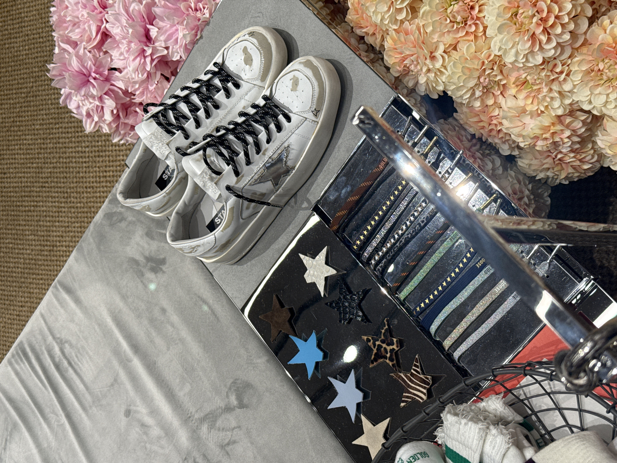 The image shows a still life on a reflective gray surface, possibly a table. A pair of white sneakers with black laces and a silver star on the side are positioned on the left. Beside them is a black container filled with various star-shaped cutouts in different patterns and colors. Stacked next to the container is a set of black cards with rows of stars. A silver metal bar crosses over the container and cards, partially obscuring them. In the lower right corner, there's a wire basket holding white socks. To the left and right of the arrangement are bunches of pink and peach-colored flowers, respectively. There is no information regarding a location in Tianhe District, China.