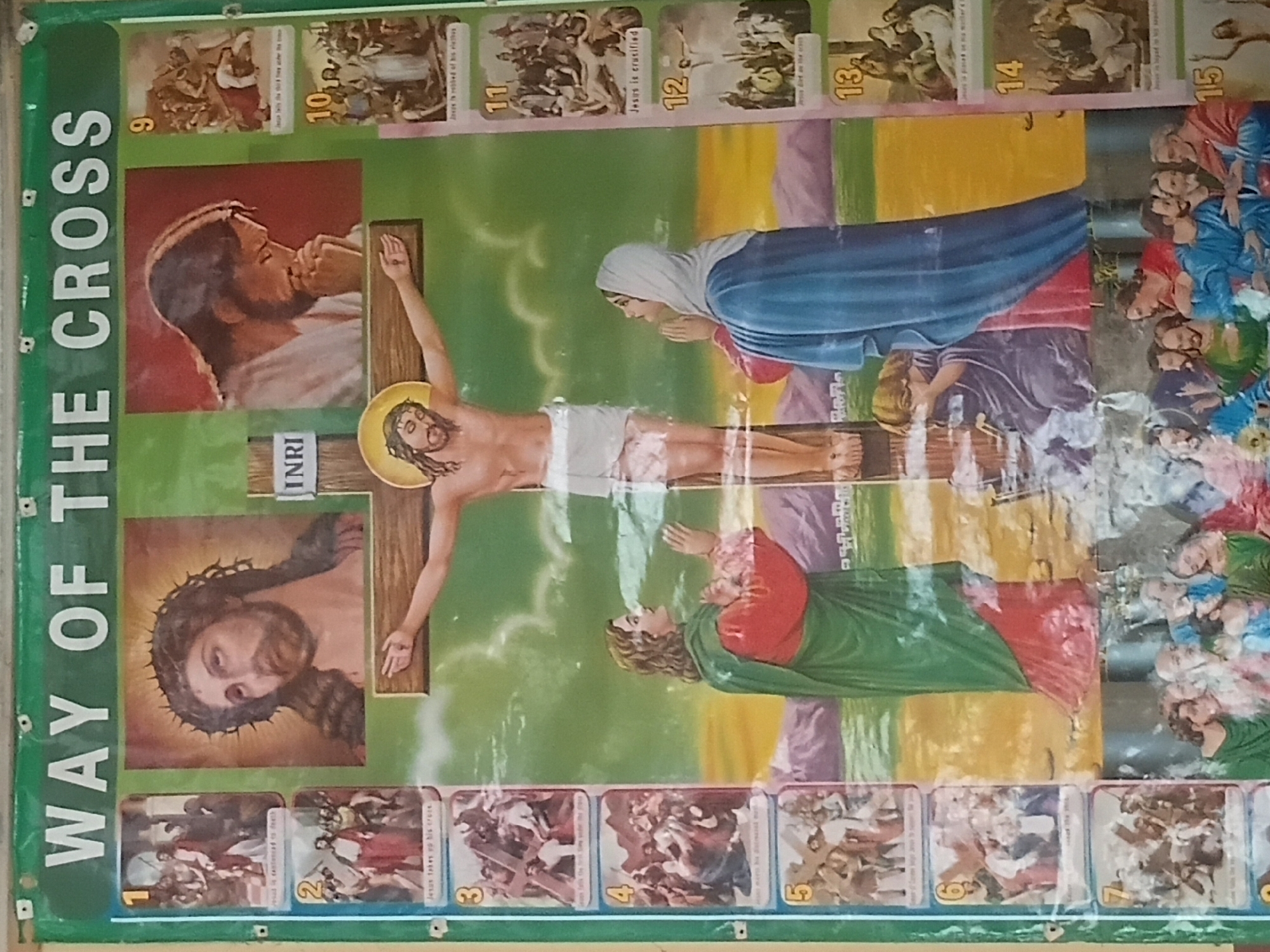 This image displays a large, vertically oriented poster depicting the "Way of the Cross," affixed to a wall in Jalingo, Nigeria. The poster has a predominant color scheme of green, yellow, and red, with various religious illustrations. At the top, a dark green banner features the white, sans-serif text "WAY OF THE CROSS." Visible metal grommets or fasteners are present along the top and left edges, securing the poster to the wall. The central and largest illustration depicts Jesus Christ crucified on a brown wooden cross, wearing a white loincloth and a crown of thorns. His arms are outstretched and nailed to the cross, with a halo behind his head. An "INRI" sign is visible at the top of the cross. Below the cross, to the viewer's left, a man in a red tunic and green cloak stands with hands raised in a gesture of lament, looking up at Jesus. To the viewer's right, a woman, identifiable as Mary, in a blue cloak and white head covering, kneels or stands with hands clasped in prayer, also gazing upwards. A third figure, partially obscured and crouched, is visible beneath Mary. The background behind the crucifixion scene features a sky transitioning from light green to a yellow-orange horizon line, with distant purple mountains. Above the main crucifixion scene, two larger portrait-style images of Jesus are displayed: one on the left shows Jesus with a crown of thorns, looking upwards with a sorrowful expression; the other on the right depicts Jesus in profile, head bowed with hands clasped in prayer, appearing contemplative. Flanking the central illustration are two columns of smaller, numbered rectangular panels, each illustrating one of the Stations of the Cross. The left column, numbered 1 through 8 (partially visible), includes scenes such as "Jesus is condemned to death," "Jesus takes up his cross," "Jesus falls the first time under the cross," "Jesus meets his afflicted mother," "Simon helps Jesus carry the cross," and "Veronica wipes the face of Jesus." The right column, numbered 9 through 14 (partially visible), includes scenes such as "Jesus falls the third time under the cross," "Jesus is stripped of his clothes," "Jesus is crucified," "Jesus dies on the cross," "Jesus is placed on his mother's lap," and "Jesus is laid in his sepulcher." A partially visible 15th panel is at the very bottom right. The lower portion of the poster, beneath the main crucifixion, is