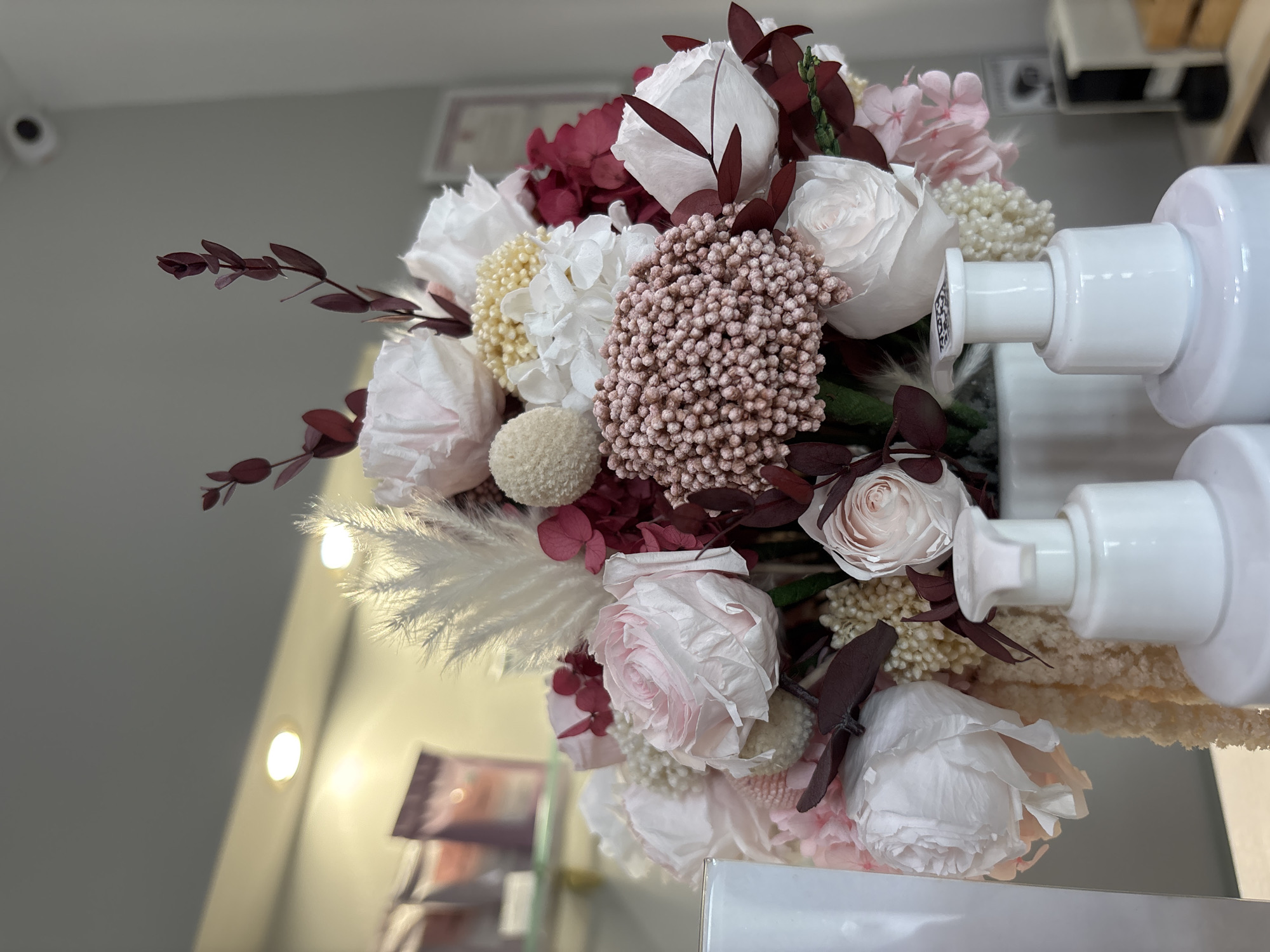 The image depicts a close-up, eye-level view of a large arrangement of dried and preserved flowers in a white vase, placed on a light-colored surface. The floral arrangement features a variety of textures and colors, including pale pink and white roses, dark red foliage, fluffy white pampas grass, and small, clustered pink and yellow blooms. In the foreground, two white pump bottles are visible, positioned in front of the vase. The background is blurred, showing a wall with recessed lighting and a security camera mounted high on the wall. The overall impression is one of a meticulously arranged display, possibly in a retail or spa setting, given the presence of the pump bottles and the professional lighting. The location is not specified.
