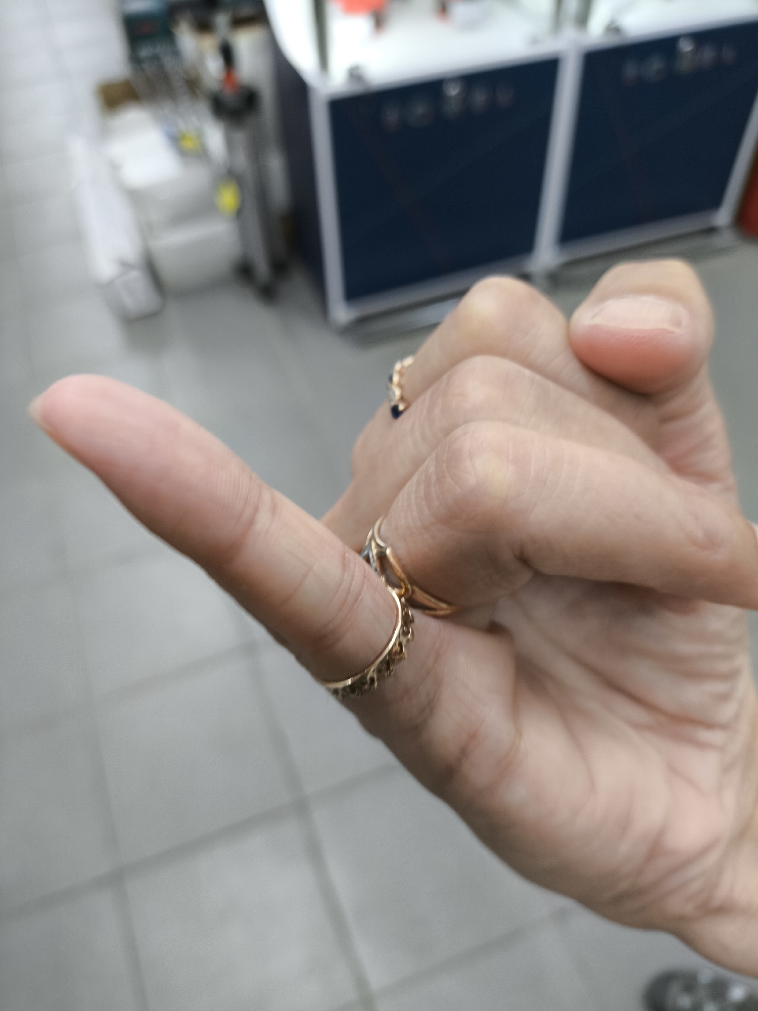 A close-up, shallow depth of field, eye-level, medium shot captures a person's left hand making a pinky promise gesture. The hand is positioned in the foreground, with the thumb extended upward and the index finger pointed outward, slightly angled away from the viewer. The pinky finger is extended, forming the gesture. The hand is adorned with several rings: a thin gold band with small blue stones is visible on the ring finger, and a more elaborate gold ring with a dangling chain and small circular charms is on the middle finger. The skin tone of the hand is light. The background is blurred, showing a tiled floor and what appears to be commercial shelving units with dark blue panels and white accents. This image does not provide any specific information to suggest it is located in the city of Solikamsk, Russia.