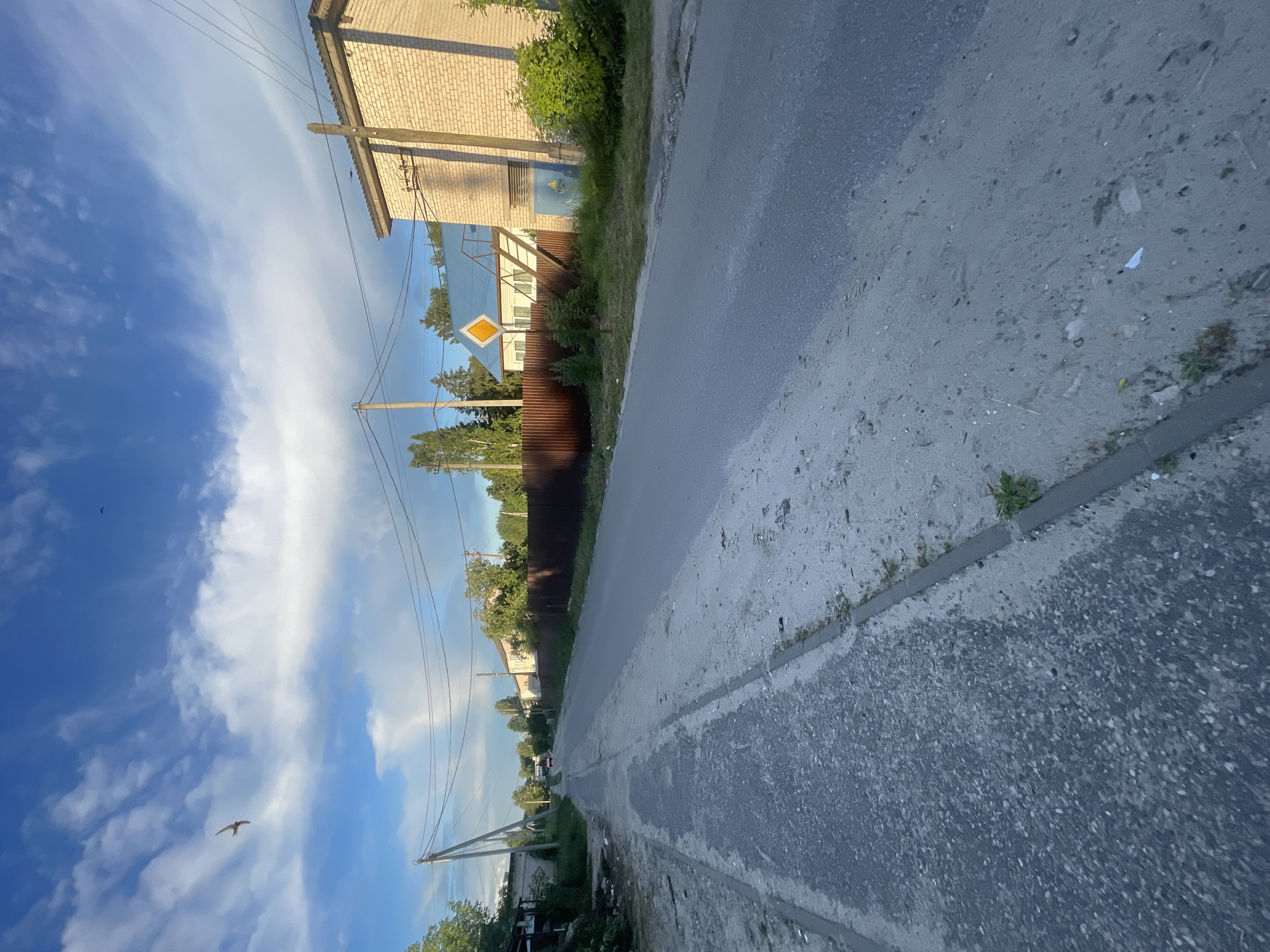 The image depicts a street scene in the settlement of Krasnye Baki, Russia. A paved road runs horizontally across the frame, with a rougher, dirt-like path along the left side, separated by a low concrete curb. Power lines are visible strung between utility poles. On the right side of the road, there is a light-colored brick building with a dark roof, and some dense green bushes in front of it. Further down the road, houses with brown fences and some trees are visible. The sky is blue with scattered white clouds, and two birds are in flight. The overall impression is a quiet, rural residential area under a partly cloudy sky.