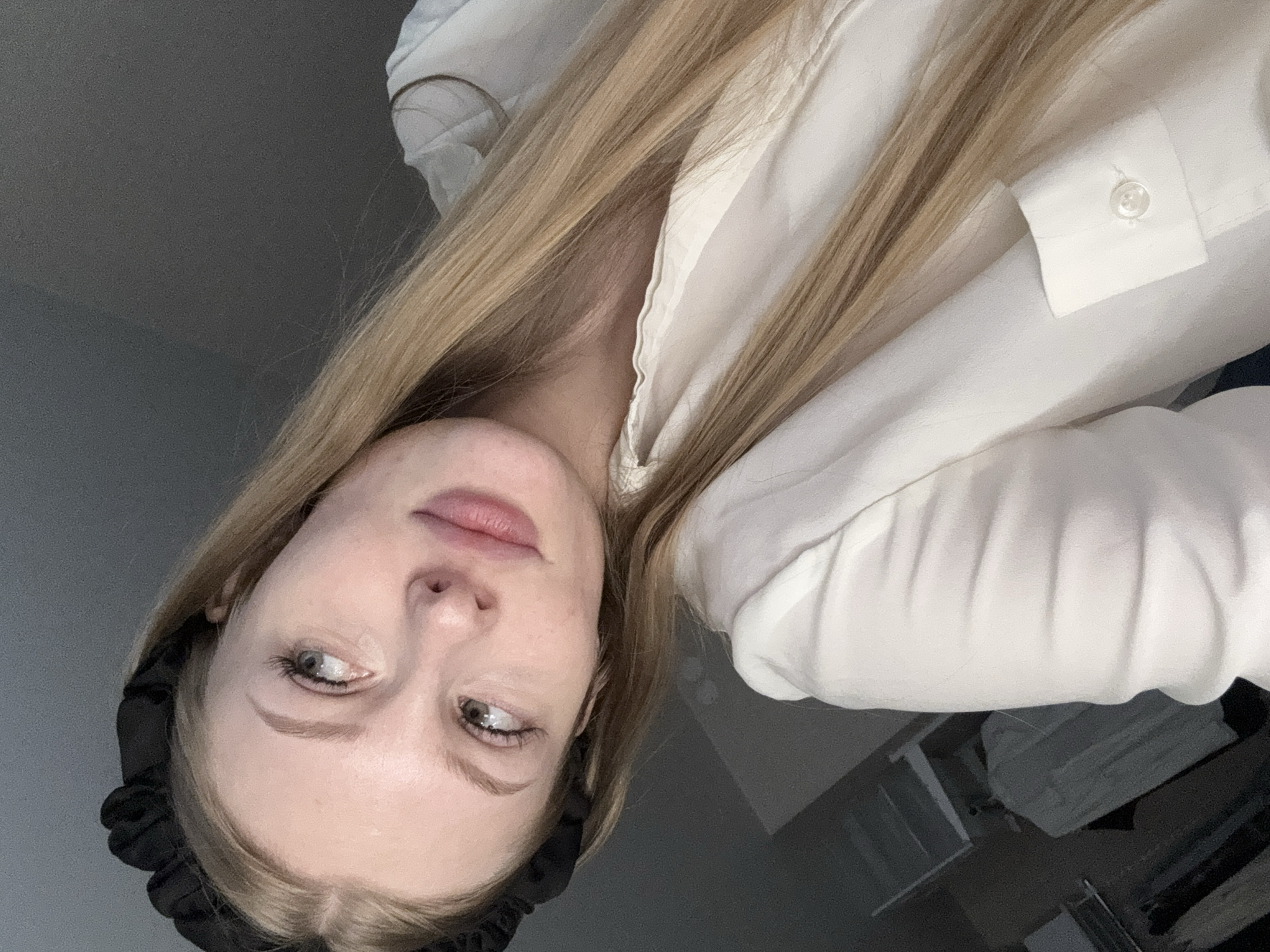 A young woman with blonde hair and a black headband is looking up and to the left, with her head tilted. She is wearing a white collared shirt. Her hair is long and falls over her shoulder and across the shirt. The background appears to be an indoor setting, possibly a bedroom, with a gray wall and some furniture visible in the lower left corner. The lighting is dim. There is no indication of the location being in the city of Perm, Russia.
