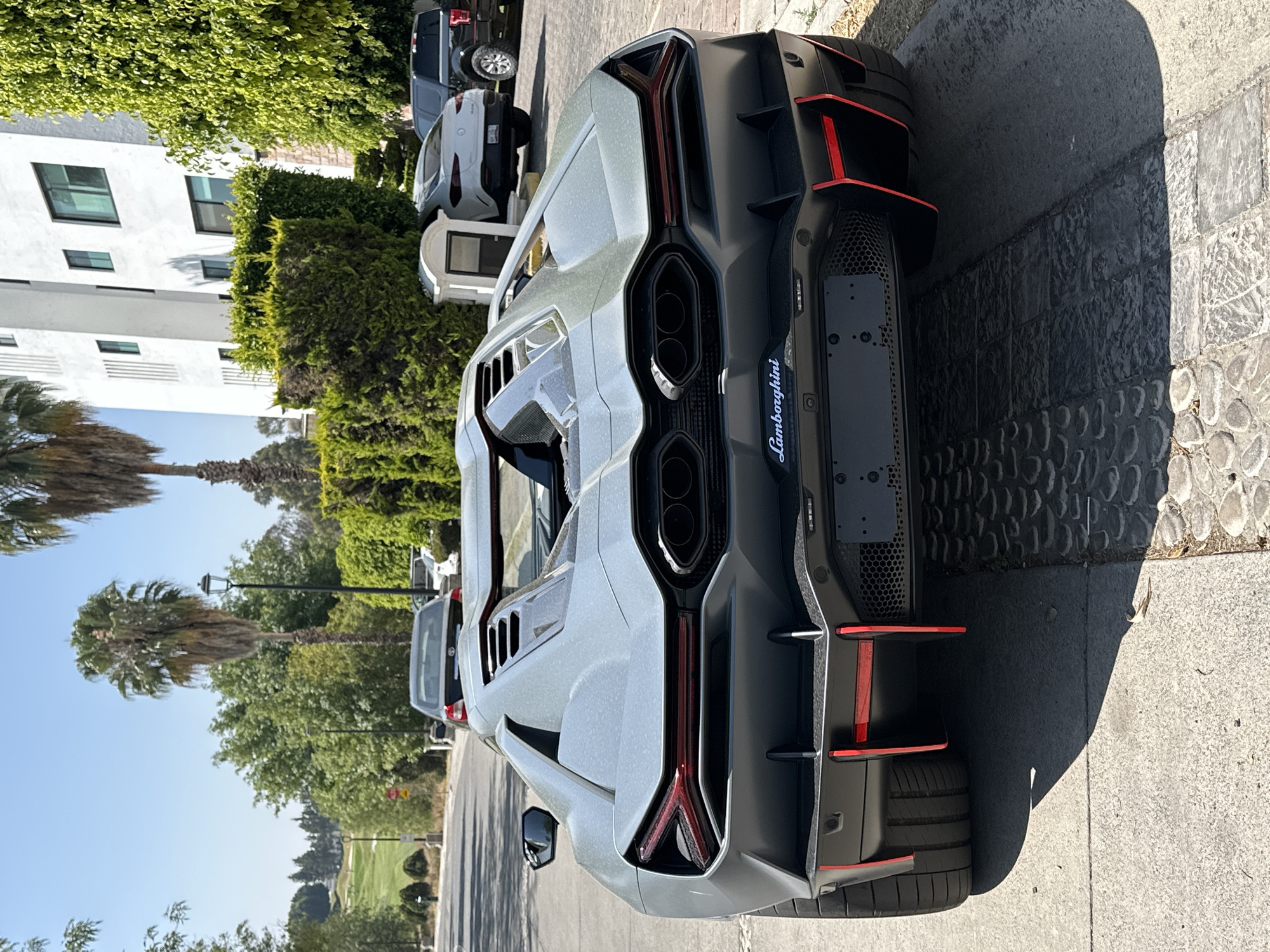 A dusty, matte grey Lamborghini Sian Roadster is parked facing away from the camera on a paved surface at the side of a road in San Bernardino Tlaxcalancingo, Mexico. The vehicle's rear displays prominent red Y-shaped taillights, four dark circular exhaust outlets, and a black diffuser accented with red vertical fins. The 'Lamborghini' script is visible on the lower bumper. The car's roof is open, revealing a dark interior. To the immediate right, the pavement transitions to a textured stone driveway. In the background, a paved road curves uphill to the left, where a silver sedan and a black SUV are visible. The surrounding environment features lush green vegetation, including hedges and tall palm trees. A multi-story white building with dark windows stands on the right. A white Mercedes-Benz GLE Coupe and a black Ford F-150 are parked further down the road on the right. In the distant background, a golf course with green fairways is visible. The scene is illuminated by strong sunlight under a clear blue sky. No people are present.