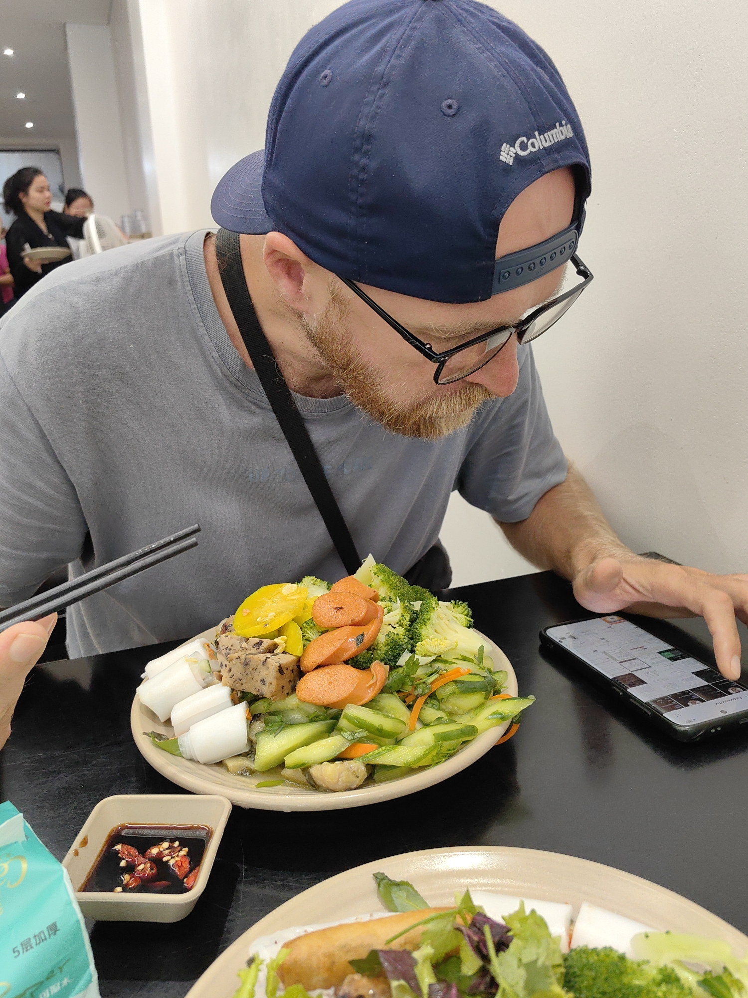 This candid photograph captures a man engrossed in his smartphone while dining in what appears to be a brightly lit restaurant or eatery in Hong Gai Ward, Vietnam.
The main subject is a man with a light brown beard and black-rimmed glasses, wearing a grey t-shirt and a dark blue Columbia baseball cap worn backward. The "Columbia" brand name and logo are clearly visible in white stitching on the back of his cap. He is looking down intently at his smartphone, which he holds in his right hand, scrolling through what seems to be a social media feed or website. A black strap, possibly from a bag or camera, is visible across his chest.
In front of him, on a dark table, rests a large beige plate piled high with a colorful and varied meal. The food includes white rolled items (possibly banh cuon or rice rolls), yellow slices (perhaps pickled vegetables), orange carrot slices, green broccoli florets, sliced cucumbers, and a block of a savory meatloaf-like item. A hand, likely belonging to another diner or the photographer, is holding black chopsticks poised near the plate. A small, rectangular dish containing dark dipping sauce with red chili flakes is also on the table. Partially visible on the far left is a green and white packet, possibly tissues, with Chinese characters "5层加厚" (5 layers thickened) and "韧" (tough/resilient) printed on it. Another plate with food, including what looks like spring rolls and greens, is in the foreground, suggesting there are at least two diners.
In the blurred background, two women are visible; one in a black top is holding a plate, possibly staff or other patrons. The setting suggests a casual, indoor dining experience during daytime. The man's focus on his phone juxtaposes with the ready meal in front of him.