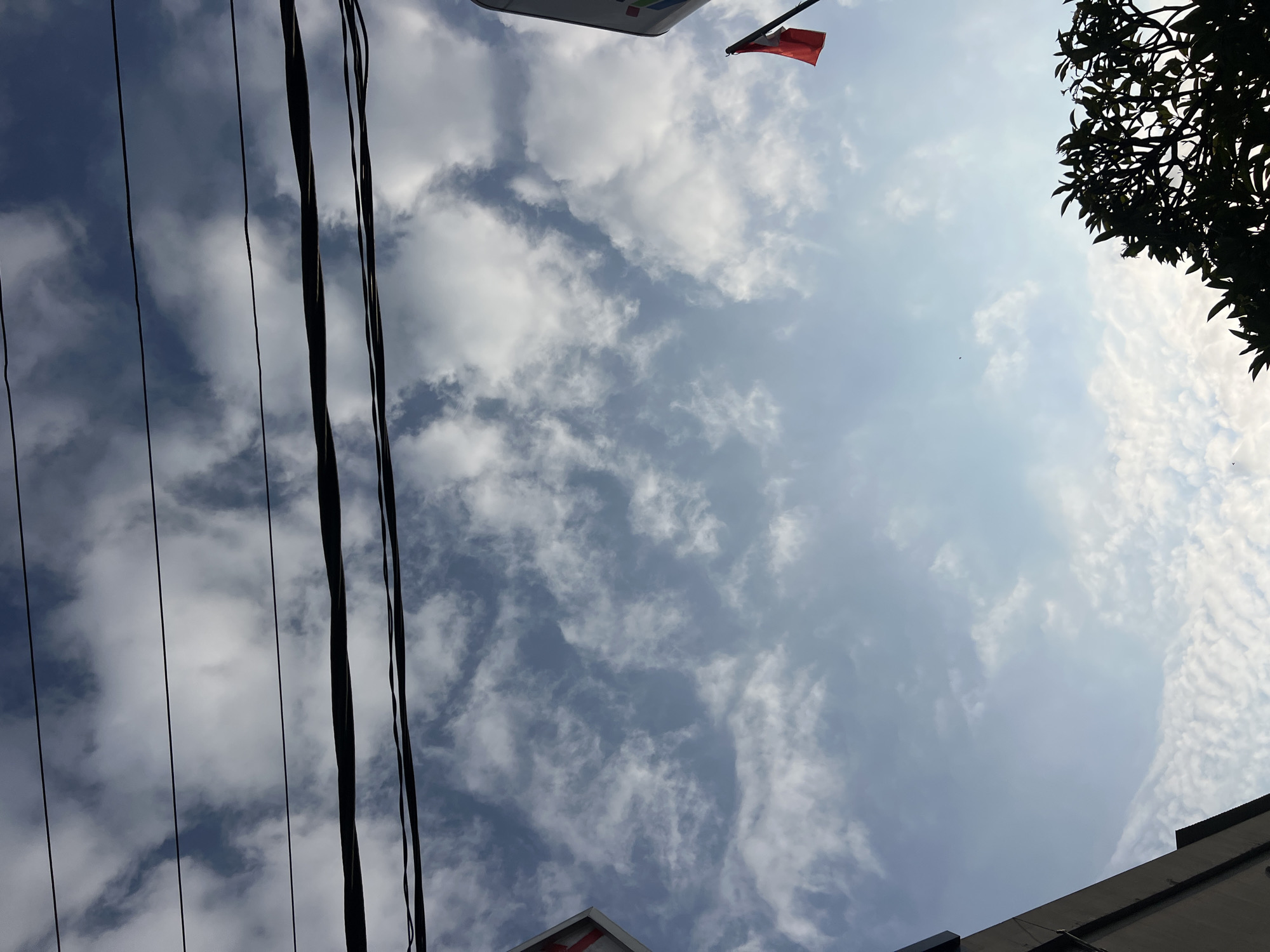 A low-angle shot captures a sky filled with scattered white clouds against a pale blue backdrop. Overhead, multiple black power lines stretch horizontally across the frame. On the right side, a small red flag flies from a pole, partially silhouetted against the sky. In the lower right corner, the dark silhouette of a leafy tree is visible. On the left side of the image, the corner of a building with a white facade and red markings is partially visible. The scene suggests an outdoor urban or semi-urban environment in Kuta, Indonesia, likely during daytime, with the overcast sky indicating potentially mild weather. There are no people or discernible actions taking place in the image.