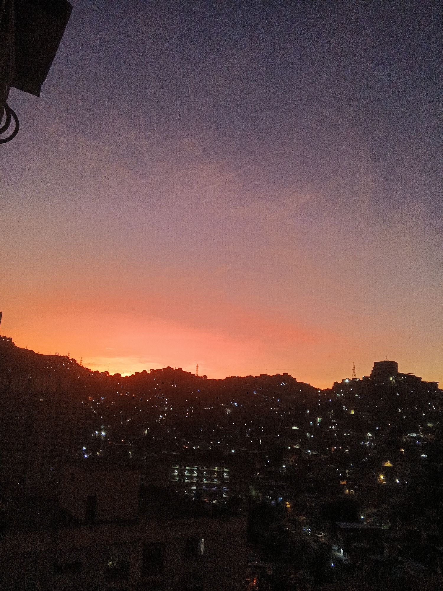 The image shows a sunset over Caracas, Venezuela. The sky is gradated from deep purple at the top to bright orange near the horizon. Silhouettes of buildings and hills are visible against the sunset. Numerous lights from buildings illuminate the hillside. In the foreground, a building is in shadow. A structure is visible in the upper left corner.