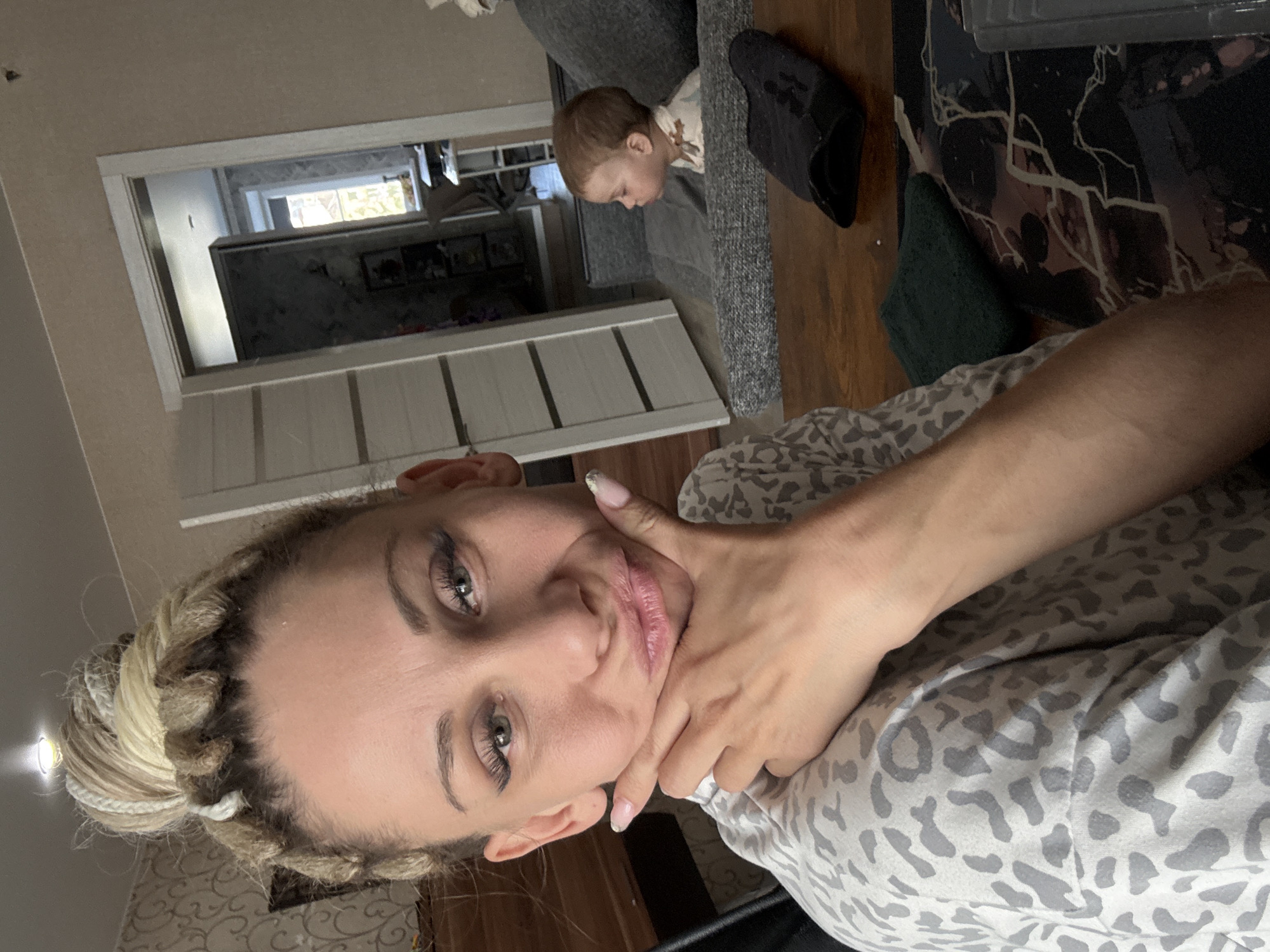 A woman with blonde braided hair tied in a bun is in the foreground, her hand supporting her chin. She is wearing a gray and white patterned shirt. In the background, a toddler is sitting on a gray sofa. A black slipper is beside the toddler. A doorway is visible in the background. The location is Rusanovo, Russia.