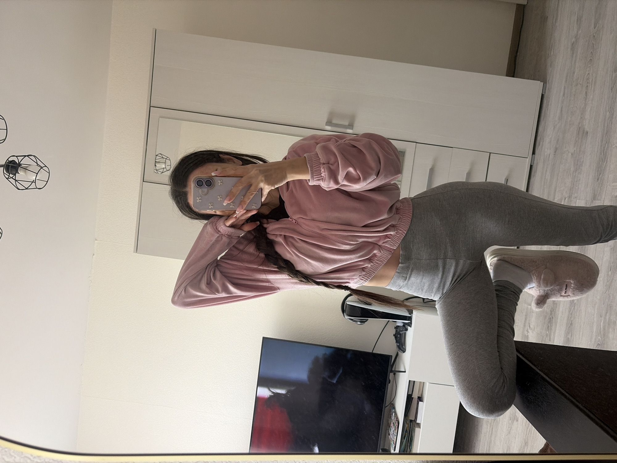 A person takes a selfie in a mirror in a domestic interior. The person is wearing a pink sweatshirt and gray leggings. The reflection shows a TV, a game console, and a white wardrobe. The location is unknown; there's no evidence to confirm it's in Saint Petersburg, Russia.