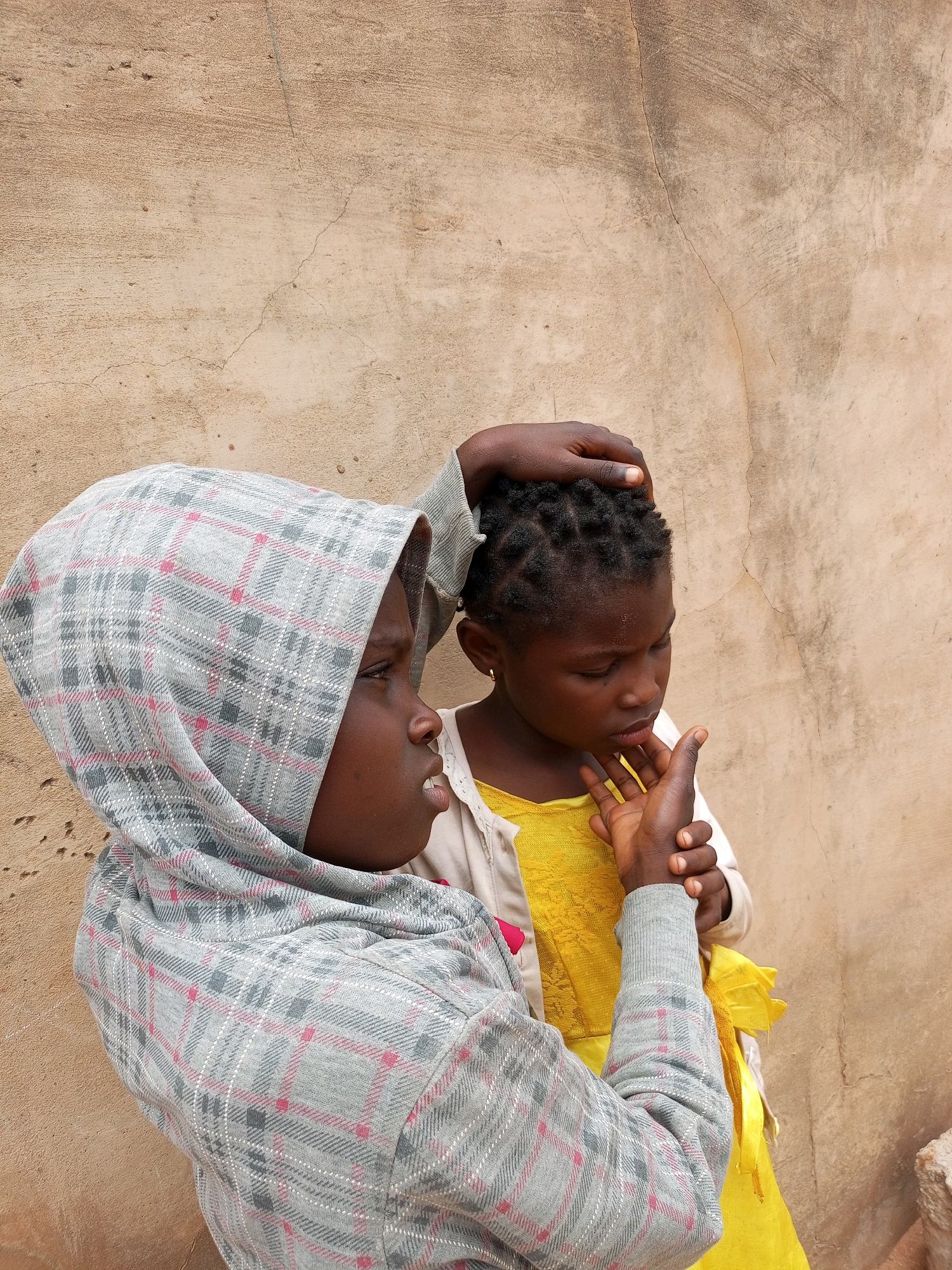 In Tempure, Nigeria, two young girls stand against a rough, tan-colored wall. The girl on the left wears a grey hooded garment with a red and white plaid pattern. She has a light skin tone and appears to be looking to her left, with her mouth slightly open. Her hand is raised, touching the head of the girl on the right. The second girl, on the right, has dark skin and is wearing a bright yellow dress with a light-colored cardigan over it. Her hair is styled in small braids, and she has a gold earring in her left ear. She is looking downwards, with her right hand held up near her face, her thumb and index finger touching. Her left hand is reaching up towards the girl on the left, touching her garment. The wall behind them has visible cracks and an uneven texture, suggesting it is an outdoor, possibly weathered, structure.