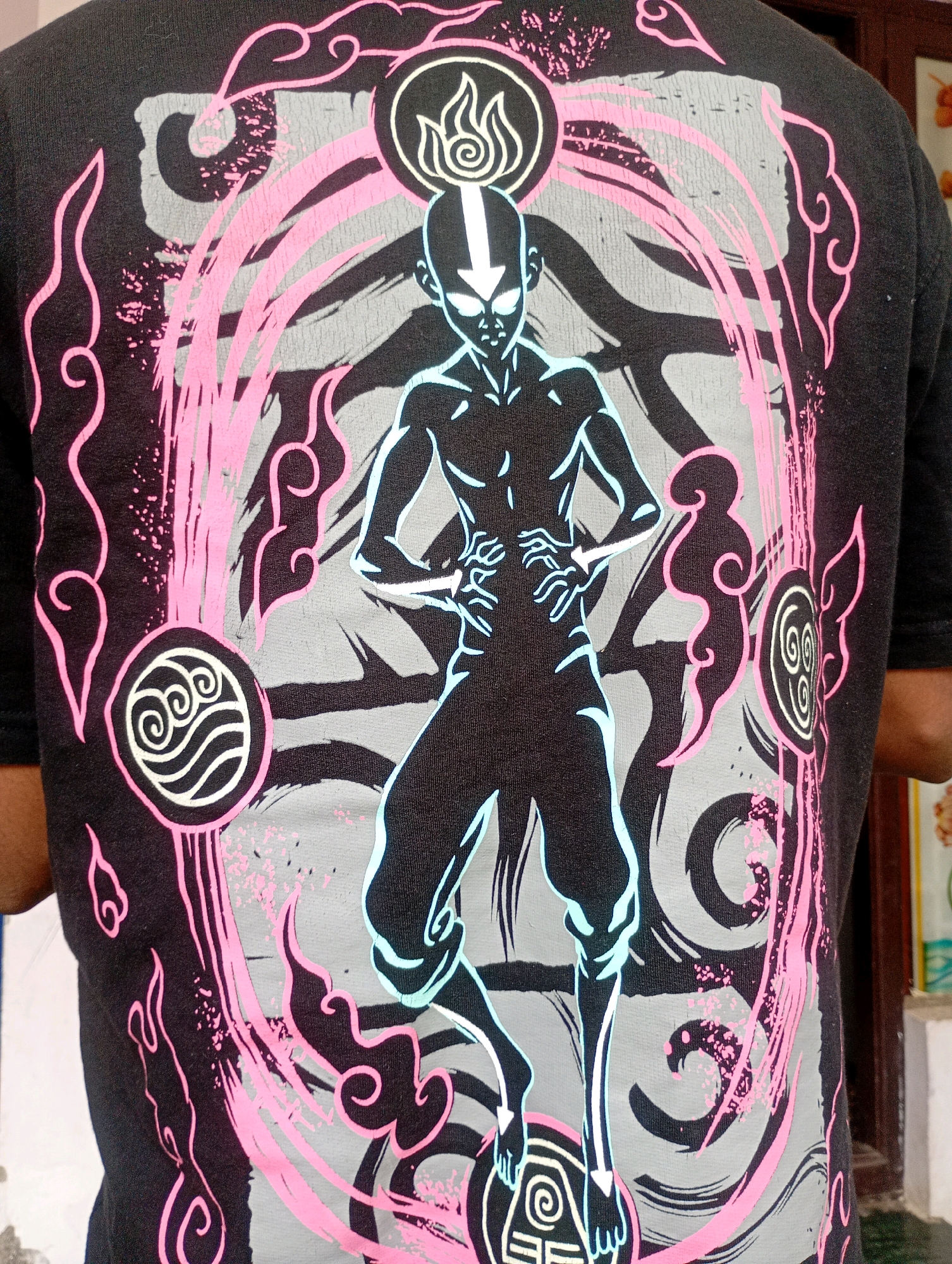This is a close-up, eye-level, full shot of the back of a person wearing a dark t-shirt with a large graphic print. The graphic features a stylized illustration of Aang from Avatar: The Last Airbender, depicted in a black silhouette with neon blue and white outlines. He is surrounded by a circular design with pink and gray elements, including swirling clouds and elemental symbols representing water, air, and fire. The t-shirt is dark gray or black. The background is out of focus, but suggests an indoor environment with a wooden surface and part of a wall visible. This image was taken in Coimbatore, India, likely in a residential or casual setting.