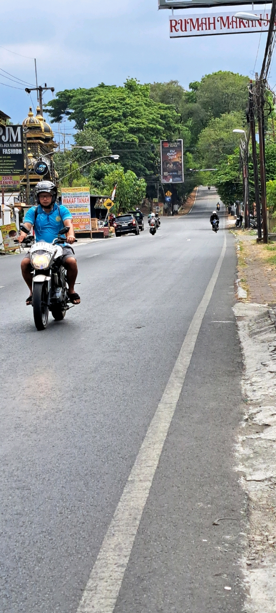 A man wearing a blue t-shirt and black helmet rides a white motorcycle down a paved road in Pandaan, Indonesia. He is wearing shorts and sandals. The road is lined with trees and poles with electrical wires. There are billboards and signs along the side of the road, some of which are in Indonesian. Other motorcycles and a black car are visible further down the road. The sky is cloudy. The scene appears to be a typical day in a suburban or rural area of Indonesia, possibly a commercial district given the presence of businesses advertised on the billboards.