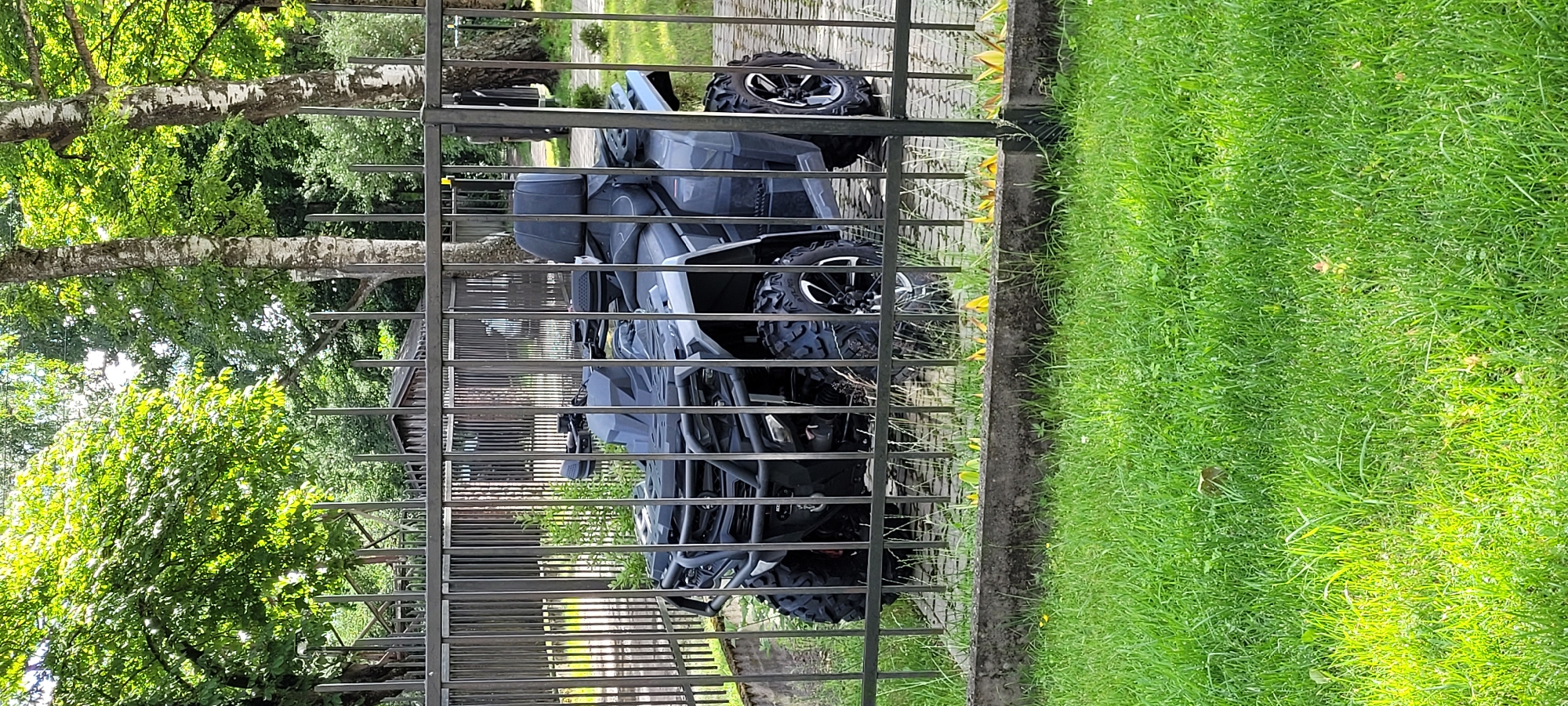 An all-terrain vehicle (ATV) is parked behind a metal fence on a paved surface. The ATV is dark gray and has a two-person seat with a backrest. It is equipped with large, knobby tires and a rugged front bumper. The surrounding environment includes green grass, trees with white bark, and some shrubbery. In the background, a small wooden structure and a taller metal structure are visible. The scene appears to be outdoors, possibly in a yard or a designated parking area within a green space. The lighting suggests it is daytime. The image quality is good, with clear details of the ATV and its surroundings. No people are present in the image. The location is not specified, but the presence of birch trees and a generally green, temperate climate could suggest a location in a northern European country like Latvia, however no specific landmarks in Riga are identifiable.