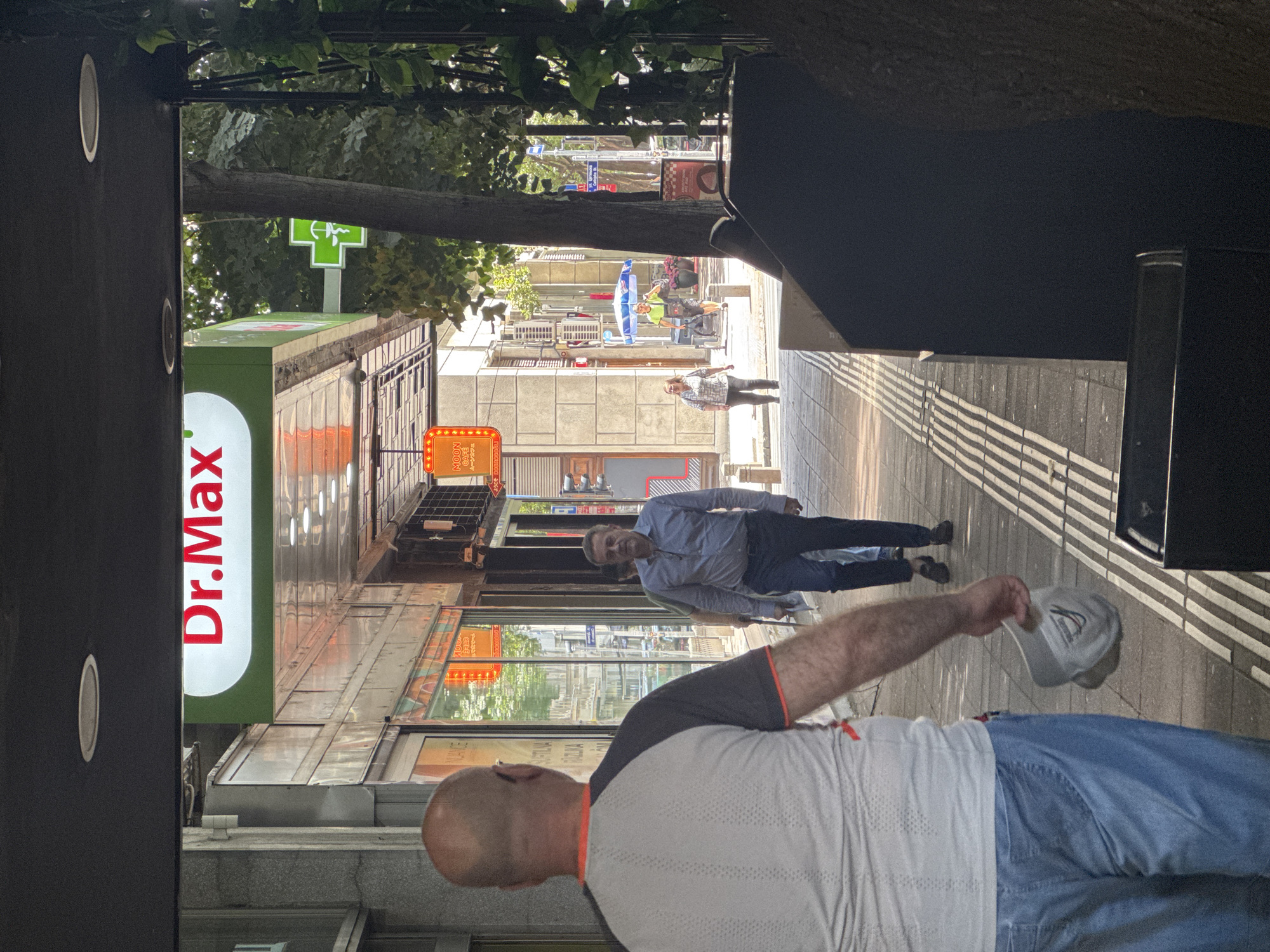 The image captures a street scene in Belgrade, Serbia, likely in a commercial area. In the foreground, a bald man wearing a grey and orange-trimmed t-shirt and blue jeans is seen from the back, walking away from the camera. He is holding a light-colored cap in his right hand.
Further down the sidewalk, a man in a light blue, long-sleeved shirt and dark pants is walking towards the camera, carrying some papers in his left hand. Behind him, another person in a lighter shirt is visible.
A prominent sign above the walkway reads "Dr. Max" in bright red letters against a white and green background. To the right of this sign, a green cross, commonly associated with pharmacies, is visible. The buildings lining the street have storefronts with reflective glass, some displaying orange illuminated signs. Trees provide shade along the sidewalk, and a dark structure, possibly a large bin or planter, occupies the lower right portion of the frame. The lighting suggests it is daytime, with sunlight casting shadows.