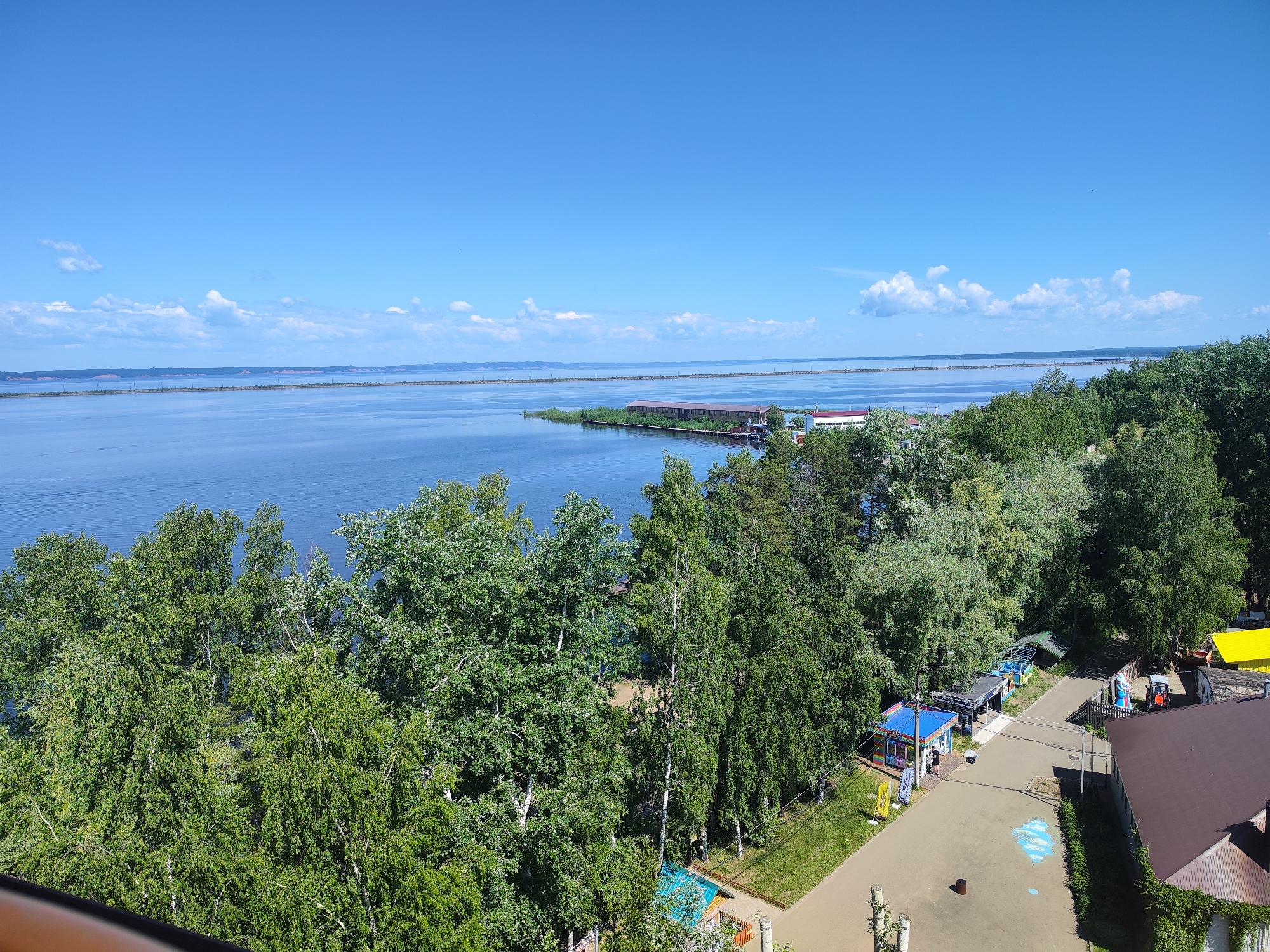 The image captures a serene waterfront scene under a bright blue sky dotted with scattered white clouds. In the foreground, lush green trees dominate the landscape, with a body of water extending to the horizon. On the right side of the image, a paved path winds through the trees, bordered by a few small buildings and structures, including a red-roofed building and a yellow-roofed structure. A few people are visible near the small buildings, engaging in unknown activities. In the distance, a long, narrow island or peninsula stretches across the water, with structures that appear to be industrial or commercial buildings. The overall atmosphere is one of a peaceful, sunny day at a recreational or coastal area.