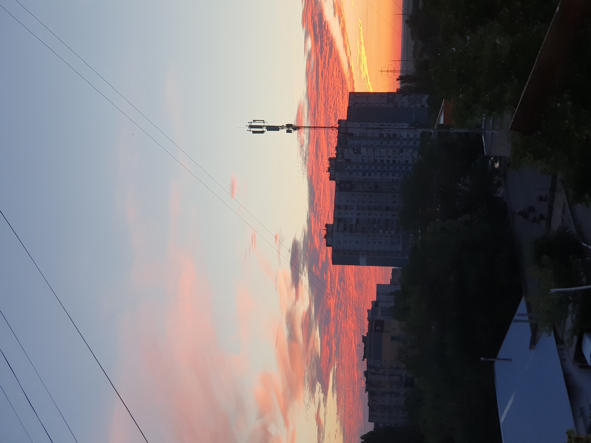 This image captures a sunset over the city of Kryvyi Rih, Ukraine. The sky is a gradient of soft blues and pinks, with dramatic streaks of vibrant orange and red clouds illuminated by the setting sun. Dominating the skyline are several apartment buildings, appearing as multi-story structures with visible windows. A prominent cell tower stands tall in the mid-ground, equipped with antennas. Silhouetted trees form a dark band across the lower portion of the scene, obscuring some of the ground-level details. In the foreground, a building with a light-colored, sloped roof is partially visible on the left. Electrical wires crisscross the upper part of the frame. The overall atmosphere is one of urban twilight.