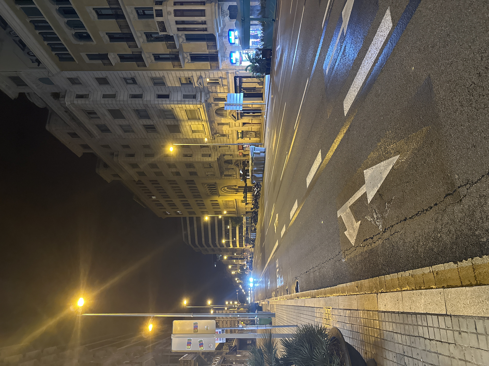 The image depicts a nighttime street scene in Barcelona, Spain, specifically on Carrer de Mallorca, near the intersection with Avinguda de Gaudí, judging by the distinctive architecture of Sagrada Familia in the background, partially visible in the distance. The street is illuminated by warm, yellow streetlights and the headlights of passing vehicles. The road is wet, reflecting the lights and creating streaks of blue and white on the asphalt. Lane markings, including arrows and dashed lines, are visible. On the left side of the road, a tiled sidewalk runs alongside several buildings. The buildings themselves are multi-story structures with balconies and windows, showing signs of urban life with some lights on in the apartments. A few parked motorcycles and scooters are visible along the sidewalk. The overall atmosphere is that of a quiet, urban night.