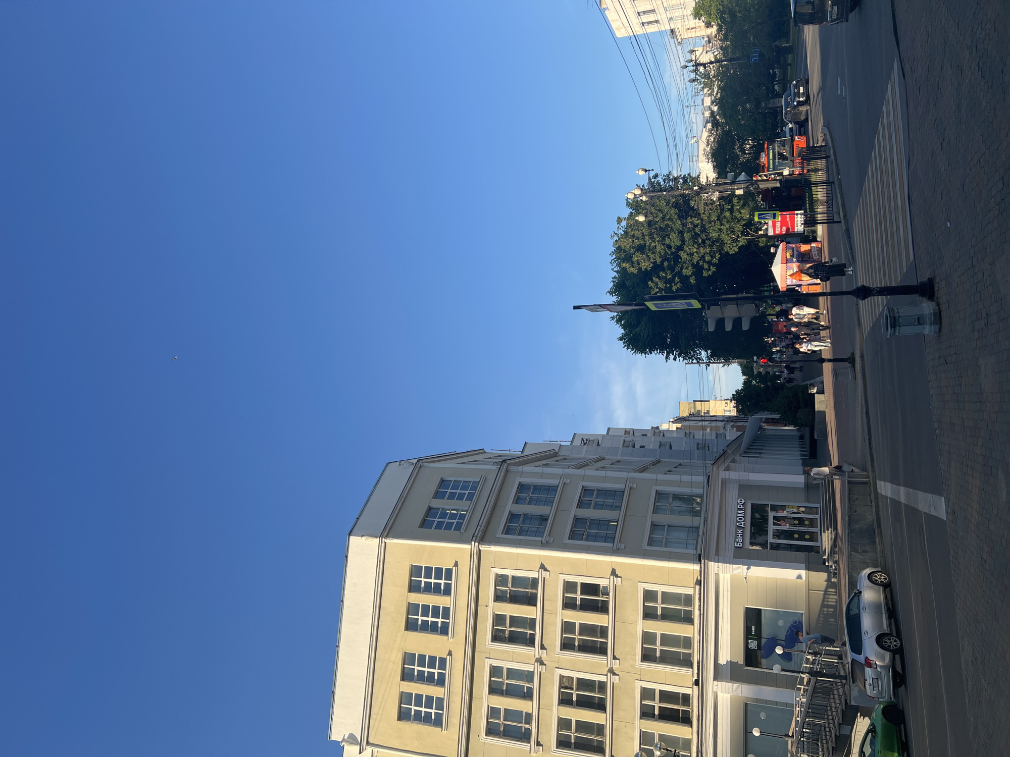 The image depicts a street scene in Khabarovsk, Russia, on a clear, sunny day with a bright blue sky. In the foreground, a pedestrian crosswalk is visible, leading into an intersection. To the left, a light yellow building with multiple windows lines the street. A silver car is parked on the side of the road.
Further into the scene, a traffic light and street signs are present. A large green tree stands prominently, casting a shadow. Various people are walking along the sidewalk and crossing the street. Buildings and more trees are visible in the background, along with power lines. One building has signage that reads "БАНК ДОМ.РФ", indicating a bank. A red truck with the word "PERTAMA" is stopped at the intersection. The architecture suggests a blend of modern and older urban design.