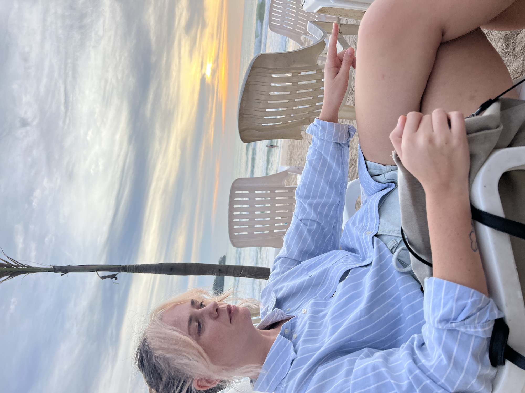 A woman with blonde hair, partially tied up, is seated in a white plastic beach chair on a sandy beach. She is wearing a light blue and white striped button-down shirt and denim shorts. Her left arm rests on a gray bag in her lap, while her right hand is extended forward, pointing to the right. The background shows a beach with several other empty white plastic chairs, the ocean with small waves, and a cloudy sky with a warm sunset glow. A palm tree is visible to the left of the woman. The scene appears to be a tranquil evening on a beach, possibly in Choeng Thale, Thailand, given the typical beach setting and vegetation.